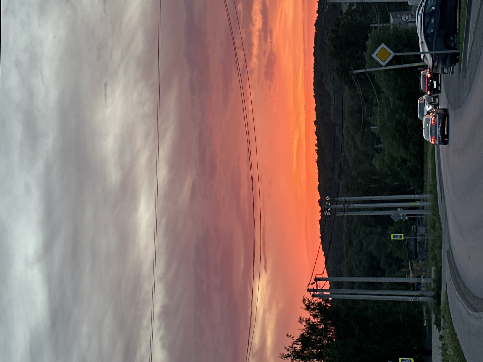 The image captures a view from Эстачи, Russia, featuring a road with several cars lined up. A yellow diamond-shaped traffic sign is visible near the cars. On the left side of the image is a dramatic sunset sky with pink and orange hues, crossed by power lines. The horizon line includes a dark, forested area. Utility poles are interspersed between the forest and the road.