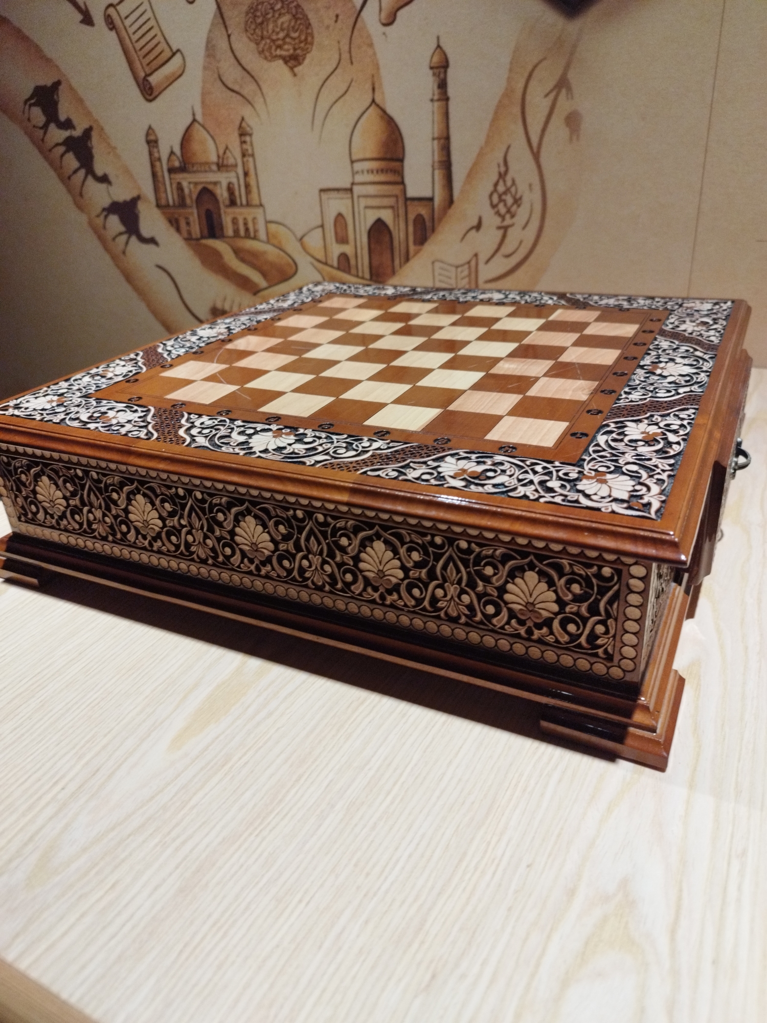 A wooden chess board with intricate decorative carvings sits on a light-colored wooden table. The chess board itself is inlaid with a checkerboard pattern of light and dark wood squares. The surrounding border of the board features detailed floral and geometric designs in contrasting wood tones. The sides of the chess board are similarly adorned with elaborate carvings. In the background, a mural or wallpaper depicts stylized architectural elements, including domes and minarets, as well as silhouetted camels and a depiction of a brain. The scene is lit from above, casting shadows on the table and the chess board.
