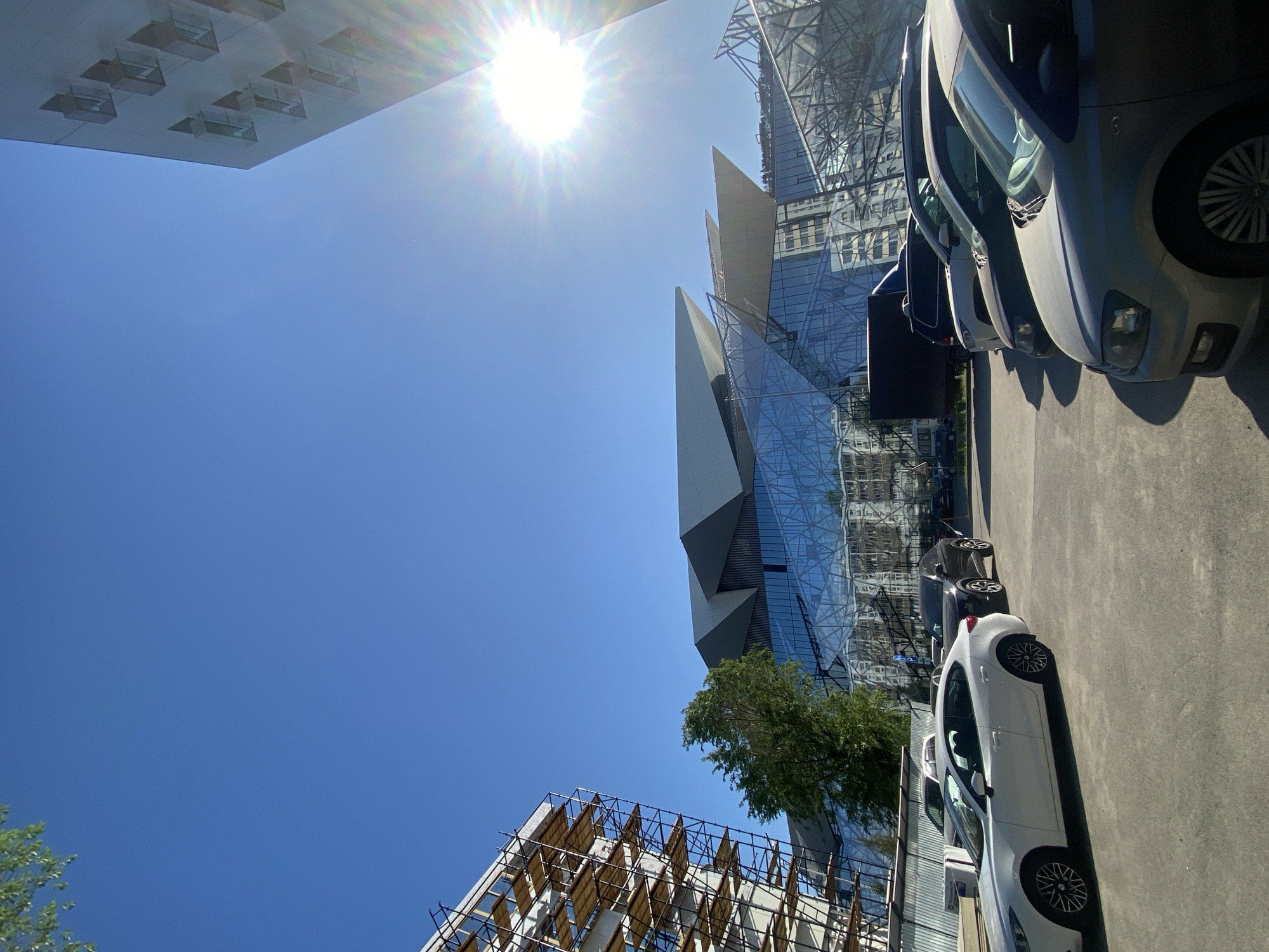 The image captures a street scene in Kazan, Russia, showcasing modern architecture juxtaposed with parked cars. A striking glass-clad building with sharp, angular lines dominates the background, reflecting the blue sky. Adjacent to it is another building under construction, partially obscured by scaffolding. A row of parked cars, including a white one in the foreground, lines the street. The sun is high in the sky, creating a bright, clear day.
