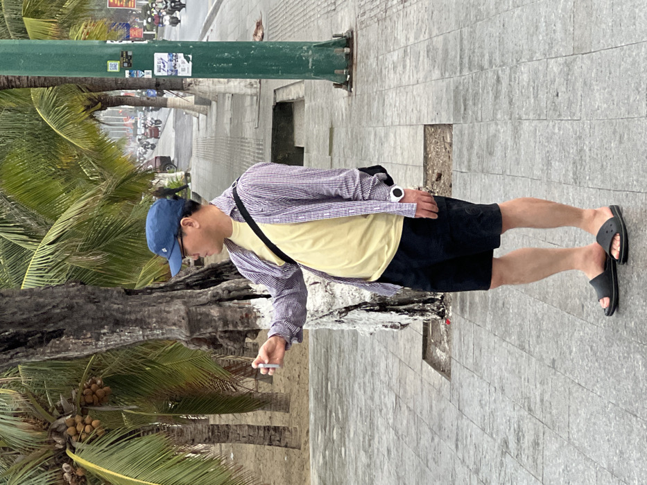 The image depicts an adult male standing on a grey tiled sidewalk in Nha Trang, Vietnam, consistent with a coastal promenade. The man, of East Asian descent, wears a blue baseball cap, eyeglasses, a light yellow t-shirt, an unbuttoned purple and white plaid shirt, black shorts, and black slide sandals. He carries a black crossbody bag and a white-faced wristwatch on his left wrist. His head is turned to the right, and he holds a white cylindrical object, possibly a vape pen or electronic cigarette, extended slightly forward in his right hand. Behind the man, a tree with rough dark bark and a white-painted base is visible, followed by a sandy area containing several palm trees with green fronds and visible clusters of coconuts. To the right of the man, a green utility pole stands, displaying multiple posters and a QR code. Beyond the sidewalk, a multi-lane road is active with vehicular traffic, primarily motorbikes, and a few cars, suggesting a busy urban environment. Further palm trees line the far side of the road. The sky appears overcast.
