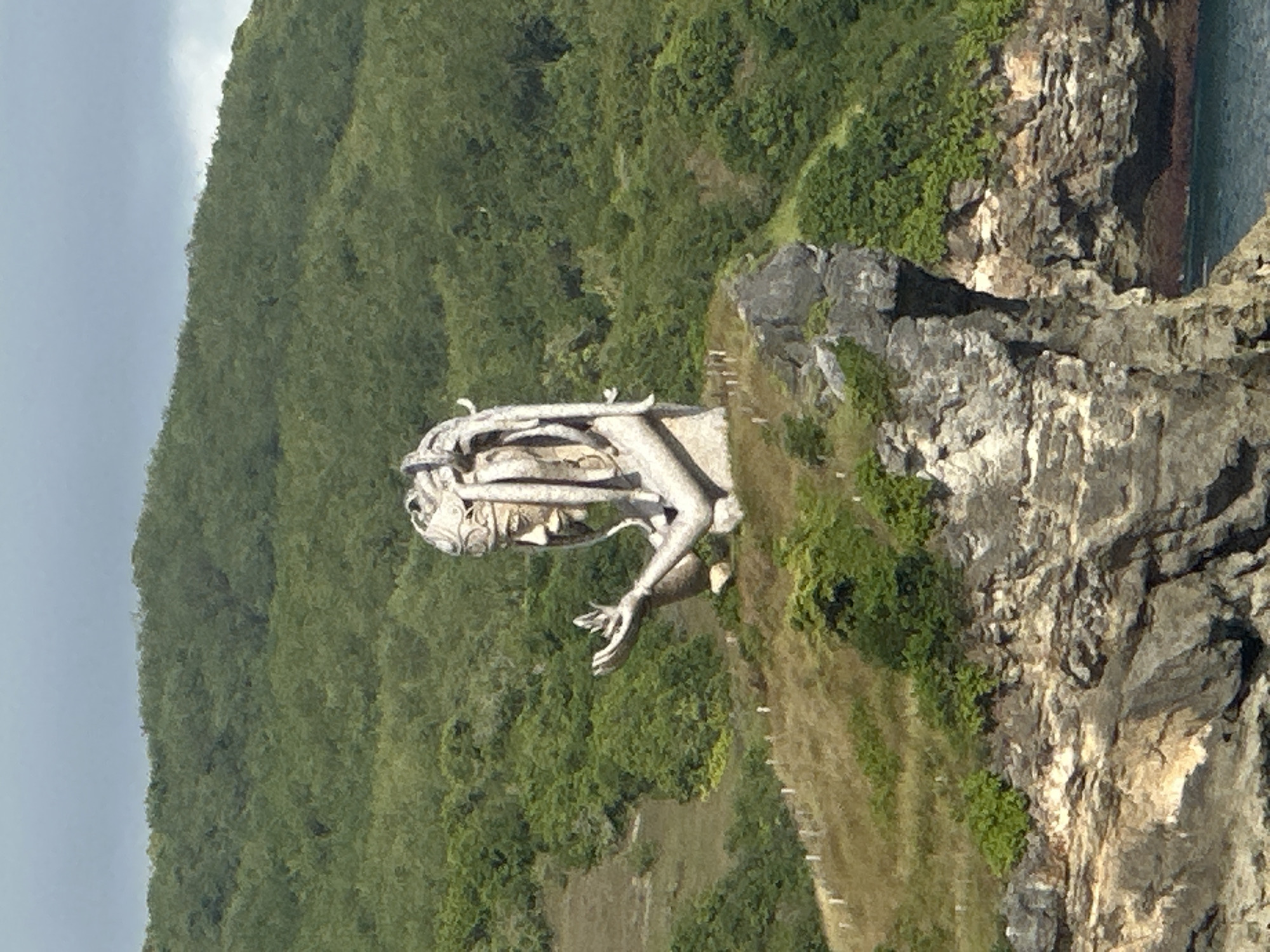 Here is a detailed description of the image:
The image shows a large, light gray statue perched on a rocky cliff in Nusa Penida, Indonesia. The statue depicts a humanoid figure, possibly a deity or mythological being, with intricate details and what appear to be decorative elements surrounding its head and shoulders. The figure has outstretched hands.
The statue is situated on a rugged, natural rock formation that forms part of the cliff edge. The cliff is covered in patches of green vegetation, transitioning from shorter grass to denser shrubbery and trees as it rises upwards.
In the background, a large, green, vegetation covered hill rises. To the right of the statue and cliff, a glimpse of the ocean is visible, indicating the coastal location. The sky appears overcast, suggesting the weather conditions.