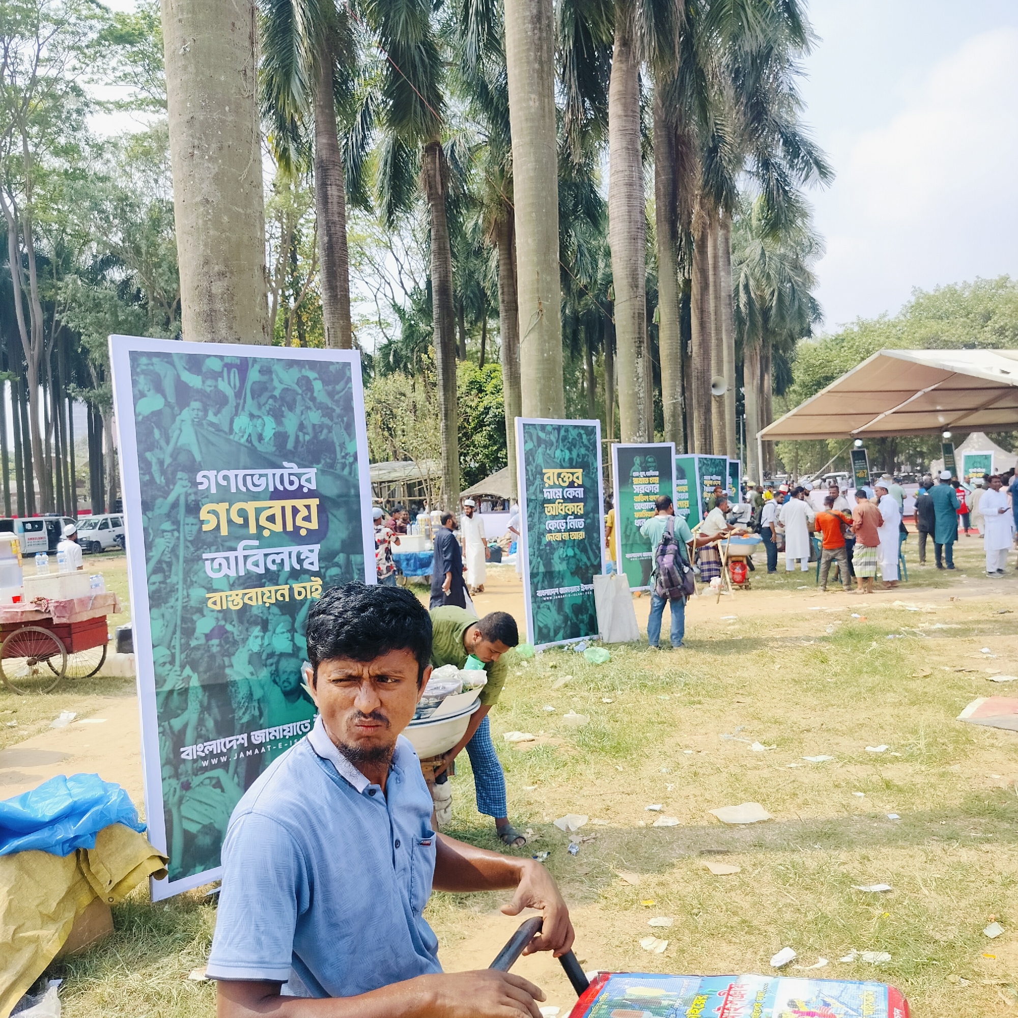 The image depicts an outdoor scene in Dhaka, Bangladesh, likely in a public park due to the presence of numerous palm trees. A man wearing a light blue polo shirt is in the foreground, possibly operating a small cart or stand, as he is holding onto what appears to be a handle. Behind him, several large signs are visible, each featuring a crowd of people and Bengali text. The signs seem to be related to political or social activism, given the assertive phrases like "গণভোটের গণরায় অবিলম্বে বাস্তবায়ন চাই" (Demand for an immediate referendum result), "রক্তের দামে কেনা অধিকার কেড়ে নিতে দেবো না আর" (We will not let the rights won at the cost of blood be snatched away again). Other people are standing and milling about further back in the scene, some wearing traditional clothing and others in modern attire. The presence of a tent-like structure in the background suggests a temporary event or gathering. The ground is covered in grass, and there appears to be some litter scattered about. A food cart and vehicles are further in the background as well.