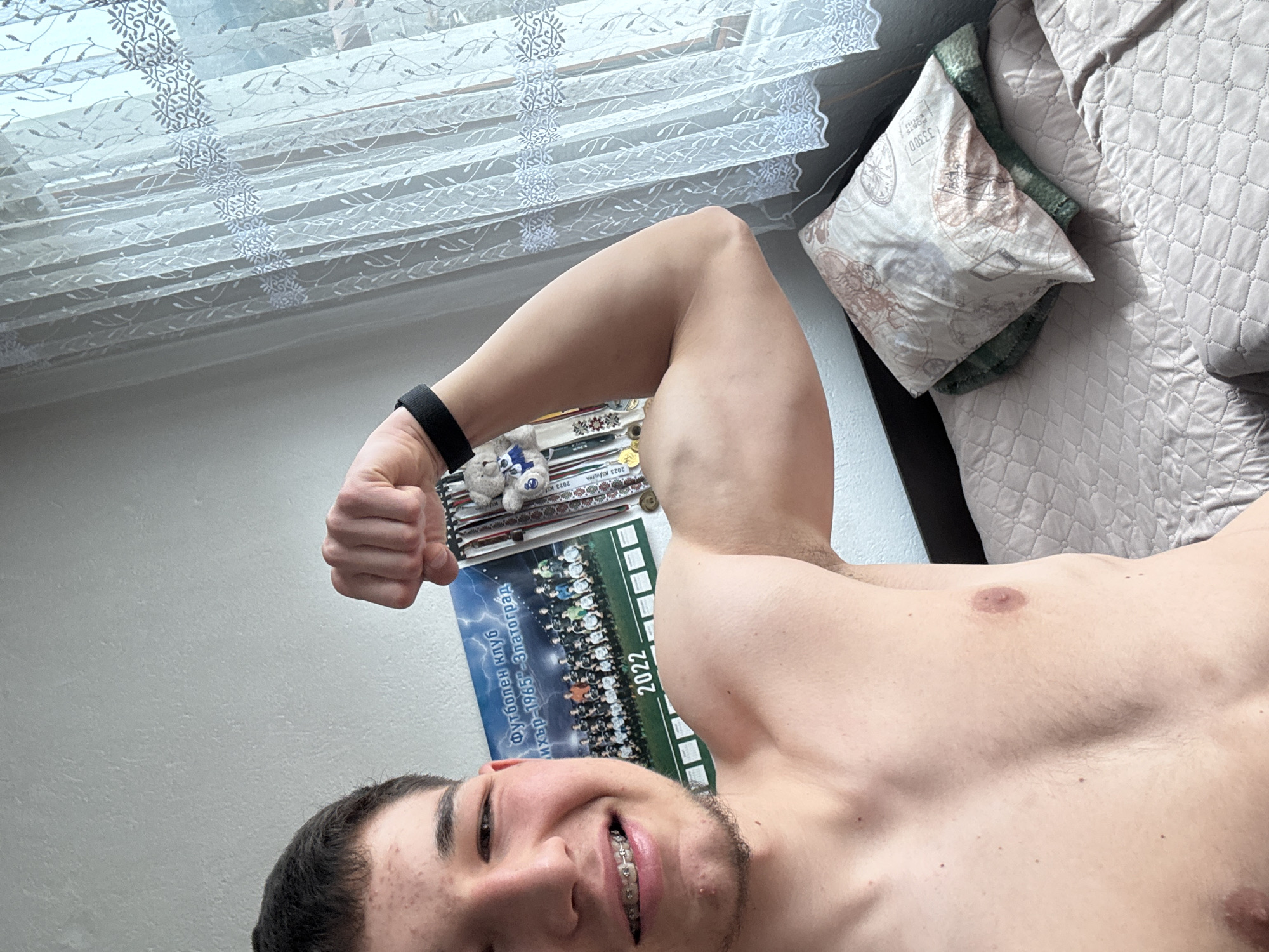 A shirtless young man with braces on his teeth is flexing his right bicep. He is wearing a black fitness tracker on his wrist. In the background is a window with lace curtains and a bed with patterned pillows. On a table are a plush toy and a poster that says "Футболен клуб "ихър-1965"-Златоград" ("Football club "Iskar-1965"-Zlatograd") and "2022", which identifies the location as Zlatograd, Bulgaria. There are also some books on the table.