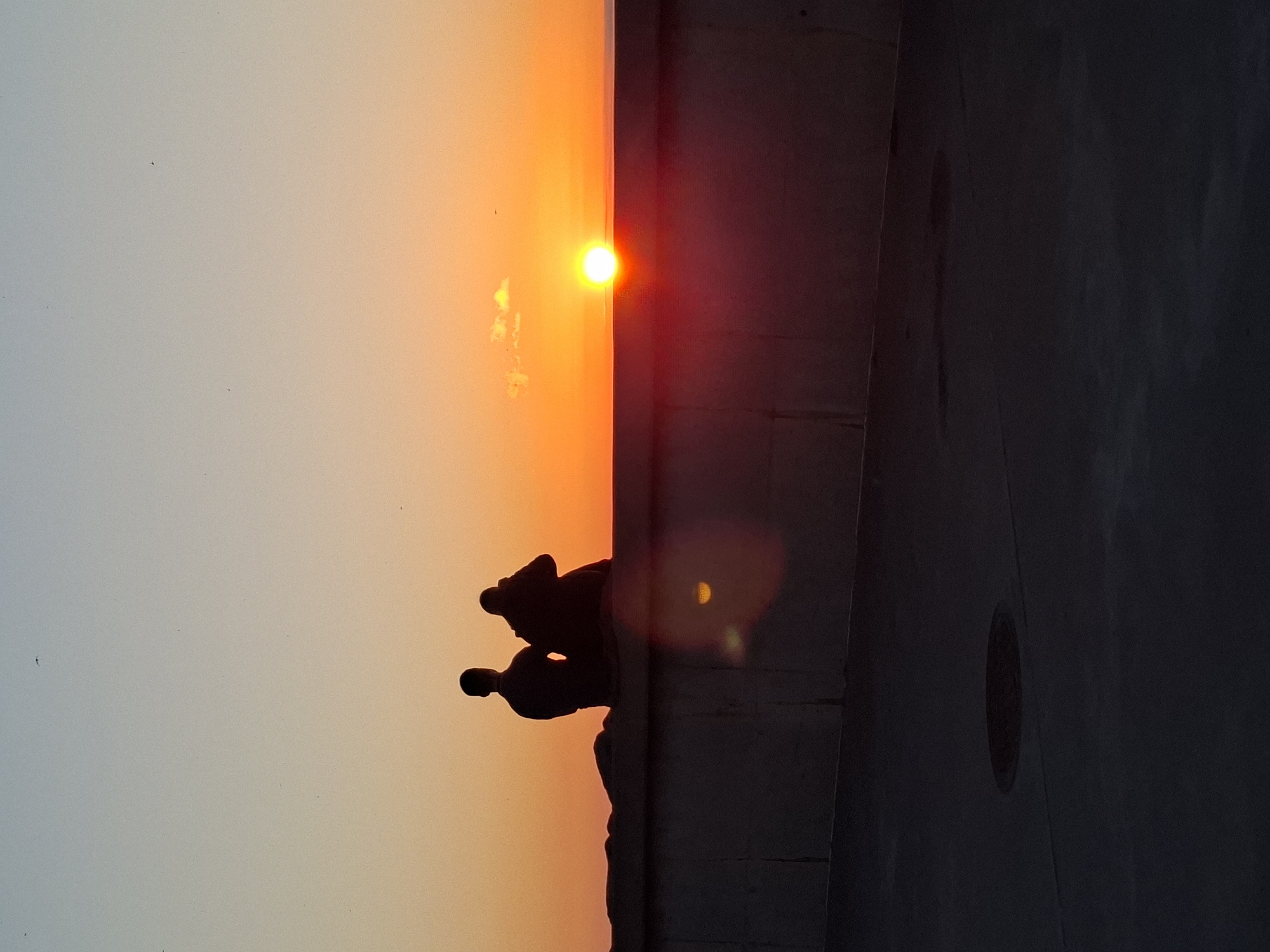This photograph captures a sunset scene in a coastal area of Tallinn, Estonia. Two human figures are observed in silhouette, seated on a dark, concrete seawall or barrier, facing towards the setting sun. The figure on the left maintains an upright posture, while the figure on the right appears to be leaning slightly forward, possibly engaging with an object such as a camera or phone, aimed towards the horizon. The sun, a brilliant white-yellow orb, is positioned just above the horizon line, dominating the scene with an intense orange and yellow glow that extends across the lower portion of the sky. The sky exhibits a gradient, transitioning from a light blue-grey at the zenith to vibrant orange and yellow hues near the horizon. Small, dark specks, likely birds or dust, are faintly visible against the upper sky. Below the concrete seawall, the foreground consists of a dark, flat paved surface, possibly a walkway, with a circular manhole cover visible in the lower-left quadrant. A distinct, circular reddish-orange lens flare is present in the lower-mid section of the image, below the seated figures, alongside a fainter reflective patch on the wall itself.