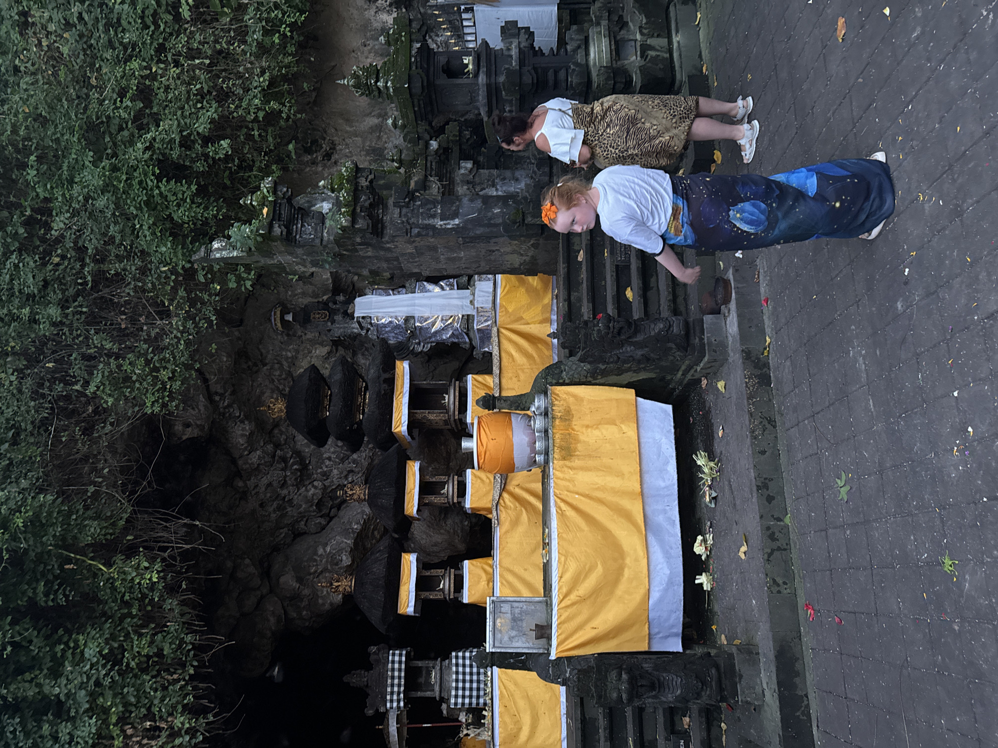 In Dawan, Indonesia, at the Goa Gajah (Elephant Cave) archaeological site, a woman and a young girl stand on a stone pathway in front of a series of offerings. The girl, facing away from the camera, wears a white t-shirt and a long, dark blue sarong patterned with blue flowers and stars. She has an orange flower in her hair. The woman, to her right and slightly behind, wears a white off-the-shoulder top and a leopard-print sarong.
The offerings are displayed on tiered stone platforms draped with yellow and white cloth. Several elaborately decorated shrines with black, tiered roofs topped with gold ornaments are visible. A large orange and white cylindrical object, possibly a donation box or offering, sits on one of the platforms, surrounded by small silver cups. In the background, the entrance to a cave is visible, partially obscured by lush green foliage cascading down the rocky hillside. The ground is paved with dark square tiles, and scattered fallen leaves and flower petals indicate a natural outdoor setting. Stone carvings of mythical creatures or guardians are present on either side of the pathways and stairs. The overall scene suggests a spiritual or cultural site.