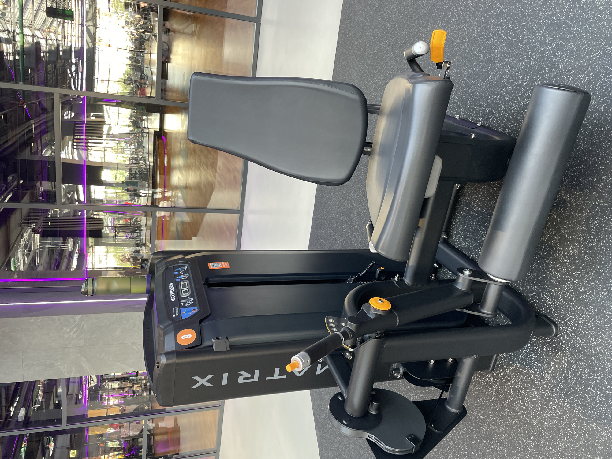 The image shows a leg extension machine with a black seat and leg pads. The machine is a Matrix brand model. The background shows the reflection of a gym and other facilities, possibly a fitness center with a modern design. The scene appears to be indoors. The city of Kazan, Russia is not evident within the image.