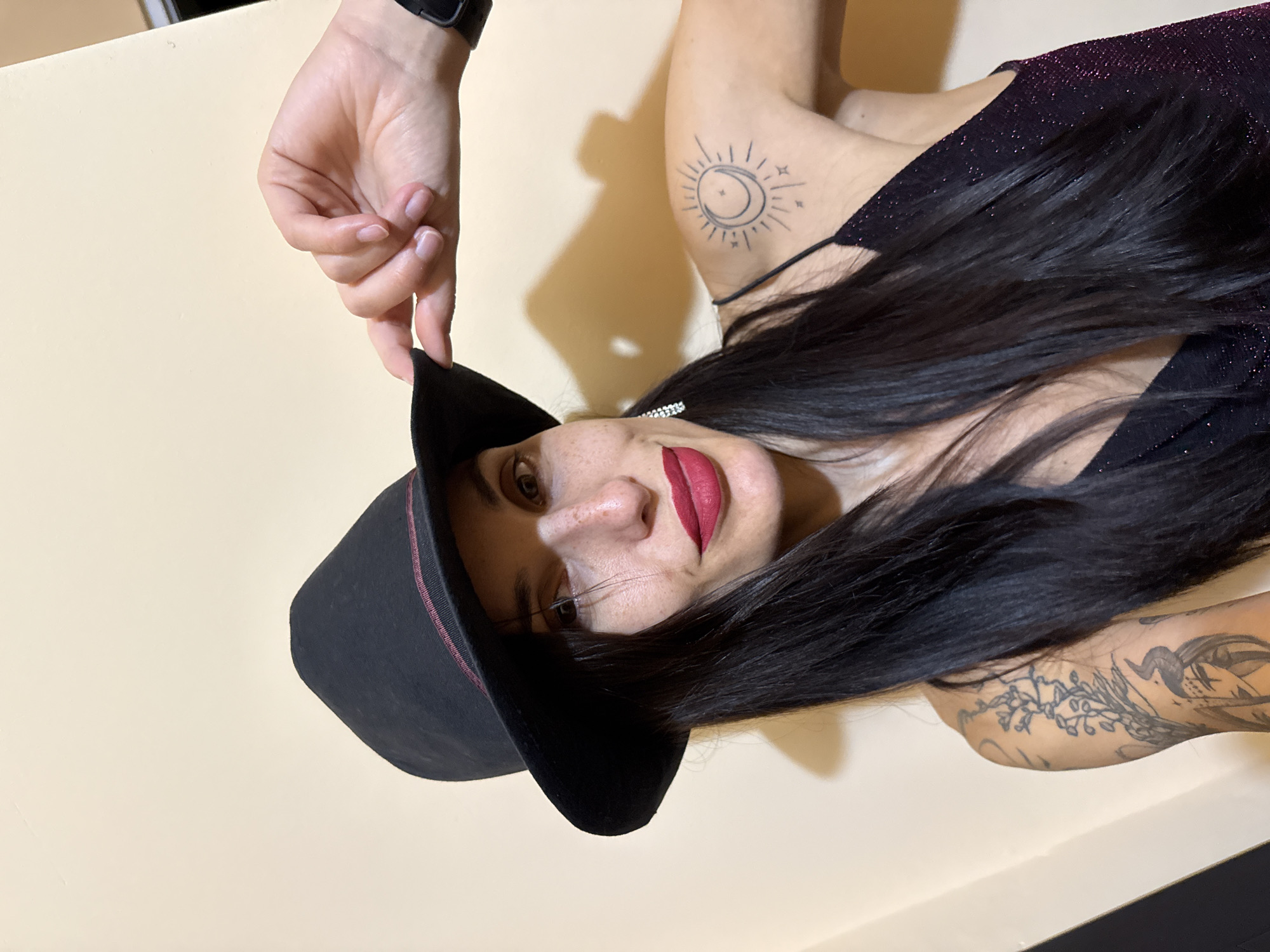 A woman with dark hair is wearing a black hat with a red trim and a sparkly black dress. She has a sun and moon tattoo on her upper arm and other tattoos on her forearm. Her hand is visible near the hat, and she is wearing a black wristband. She is wearing red lipstick. The background is a plain light yellow. There is no indication of the city of Pleven, Bulgaria, or any other specific location.