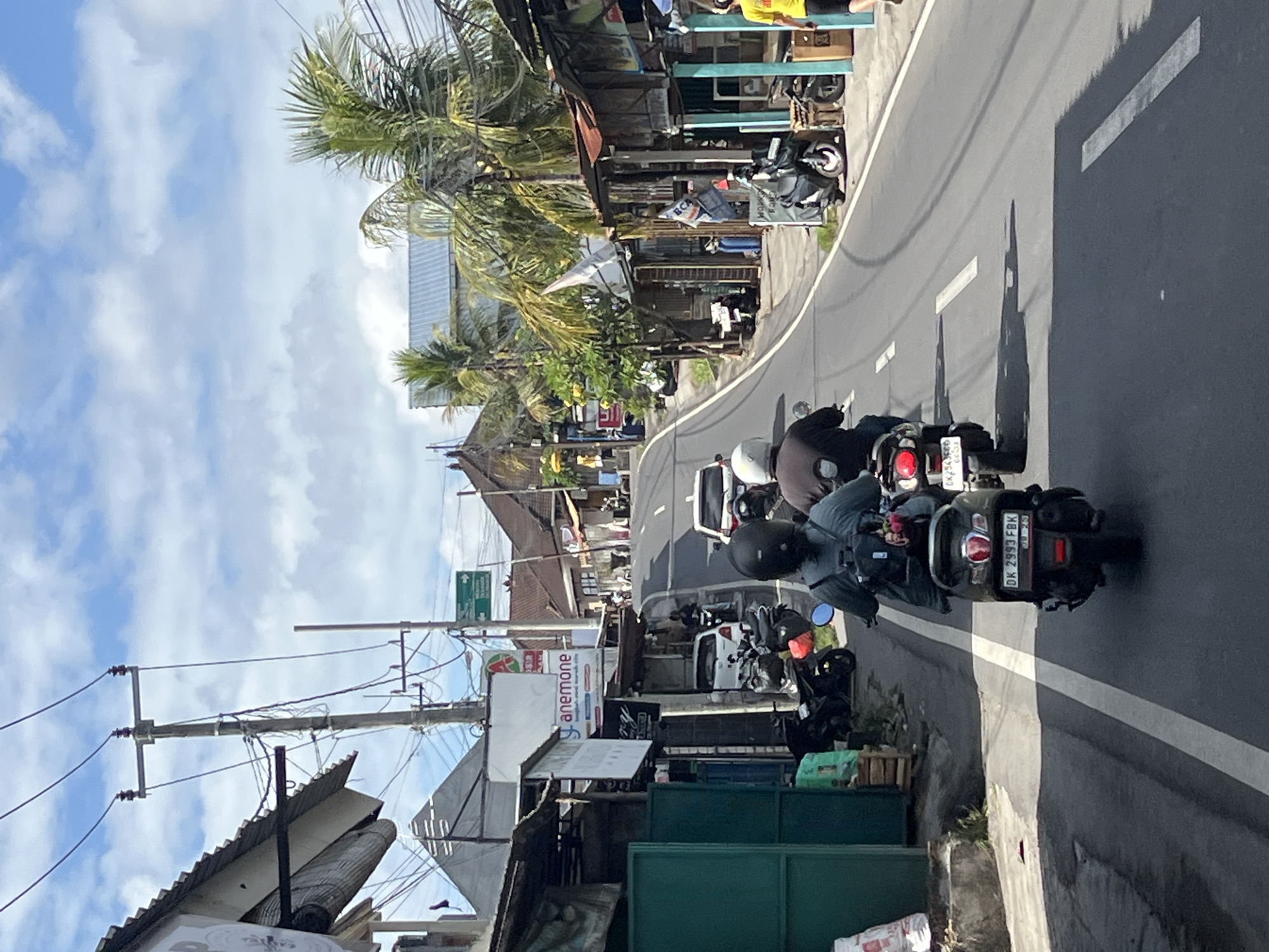 A street scene in North Kuta, Indonesia, shows traffic on a paved road. In the foreground, three motorcycles are visible. The first motorcycle, a dark color, is being ridden by a person wearing a black helmet and a dark top, facing away from the camera. The second motorcycle, also dark, is occupied by a person with a white helmet and a dark top. The third motorcycle, a lighter color, has a rider with a white helmet and a dark top. The road is bordered by buildings on both sides, with some appearing to be shops or small businesses. Palm trees are interspersed with the buildings. Overhead, power lines crisscross the sky, which is partly cloudy. The general atmosphere suggests a warm, sunny day.