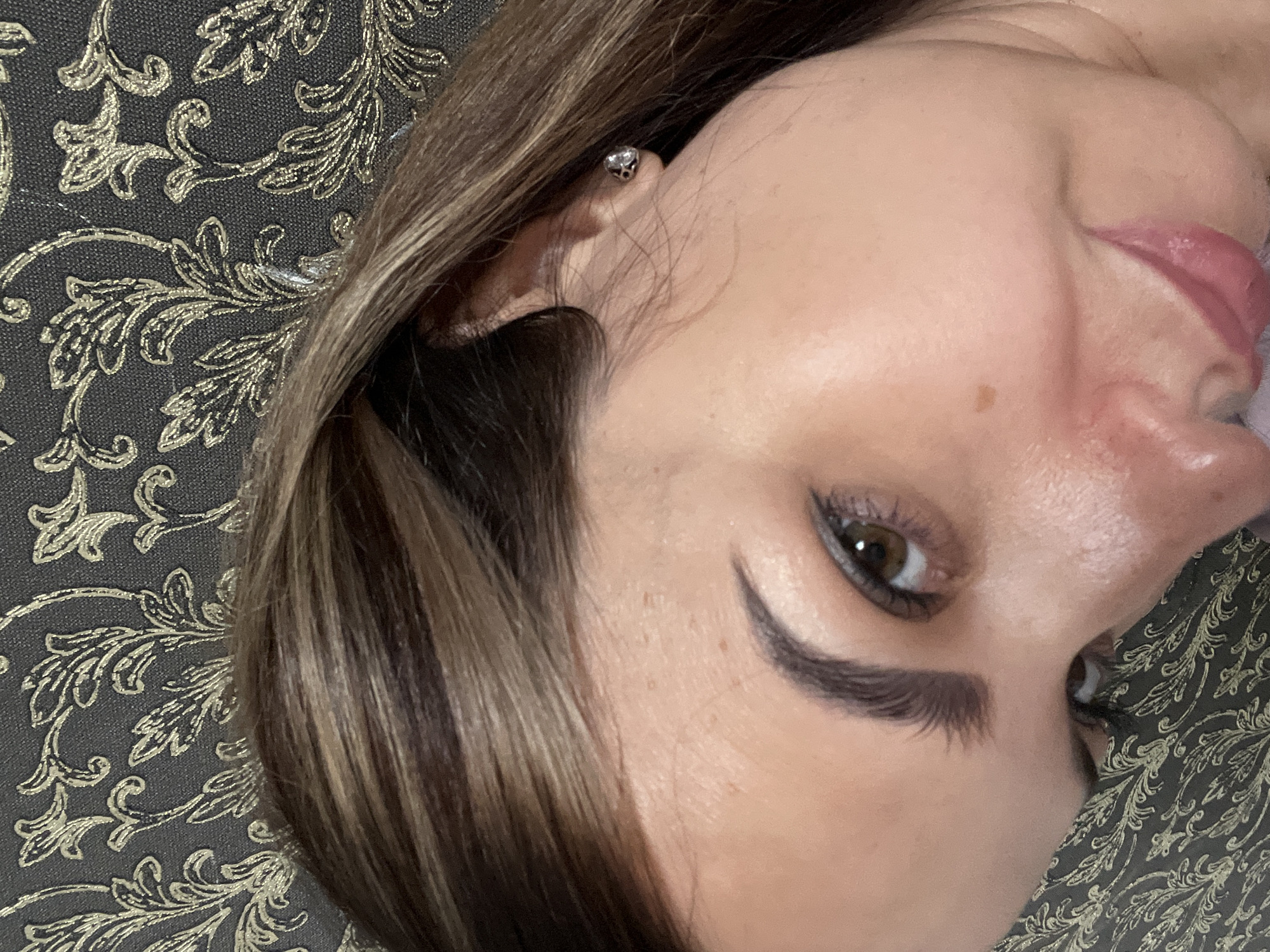 A close-up, slightly angled selfie of a woman's face. Her brown hair, with lighter streaks, is brushed back from her face. She has brown eyes, dark eyebrows, and is wearing a small, sparkly earring in her visible ear. Her lips are painted a rosy pink color. She is leaning against a dark, textured background with a gold floral pattern. The lighting is soft, creating a gentle glow on her skin. The image is sharp and in focus.