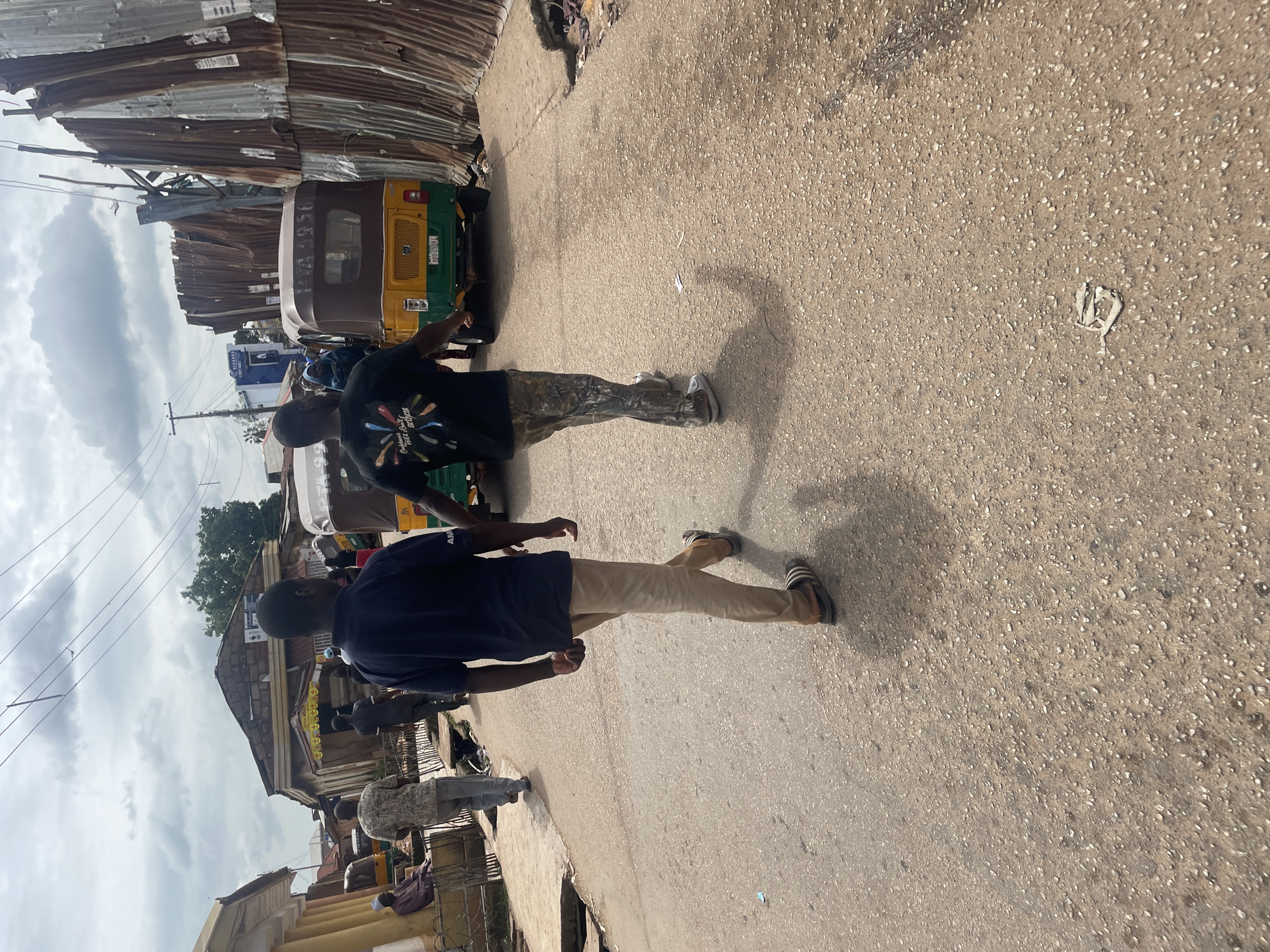 This image depicts a street scene in Gura Top, Nigeria, under a cloudy sky. Two men are walking away from the camera on a paved road. The man in the foreground on the left wears a dark blue t-shirt and light brown pants, with Adidas sandals. The man to his right wears a black t-shirt with a colorful graphic and camouflage pants with white sneakers. Several auto-rickshaws, known locally as Keke Napep, are present on the street. On the left side of the road, a man in a patterned shirt and grey pants walks away from the camera, and another person, partially visible, sits on a bench. The right side of the road is lined with buildings constructed with corrugated metal and some with a stone facade. The road surface is asphalt with scattered gravel. Trash is visible on the road and the roadside. The overall impression is of a bustling, everyday street in a Nigerian city.