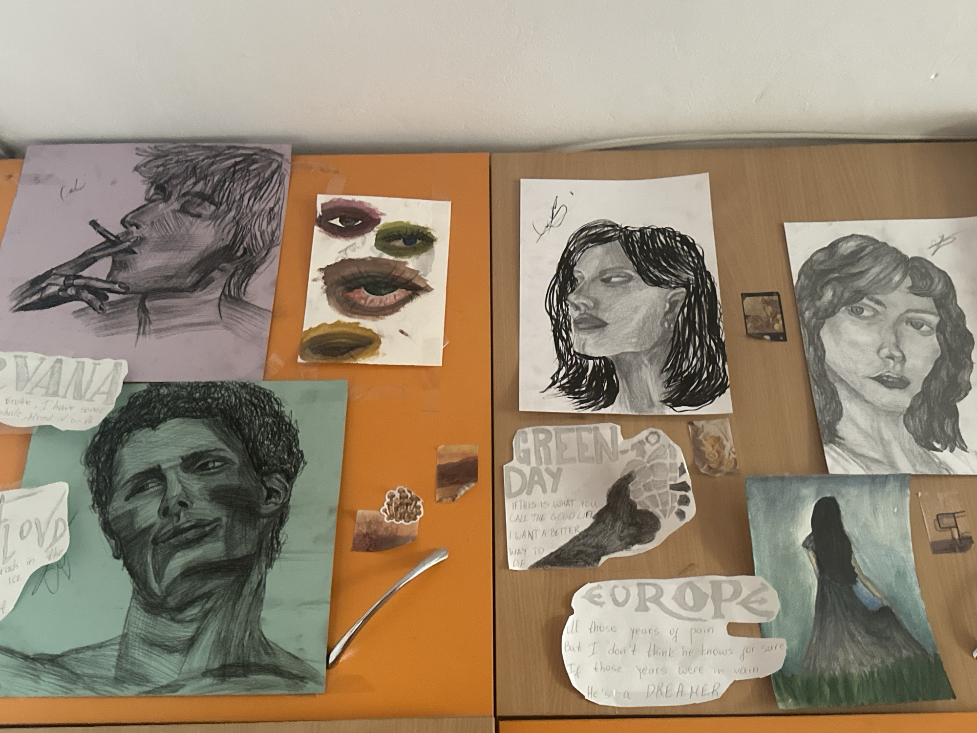 The image shows a close-up, eye-level shot of a wall collage displaying several pieces of art and text. The collage is mounted on an orange surface, which appears to be a cabinet or desk. The artwork includes charcoal sketches of portraits, a watercolor painting of a person in a dress, and a small painting of eyes. Text fragments are also visible, some appearing to be lyrics or quotes. The overall impression is of a personal collection of creative work. There are no visible indications of Plovdiv, Bulgaria, or any specific city.