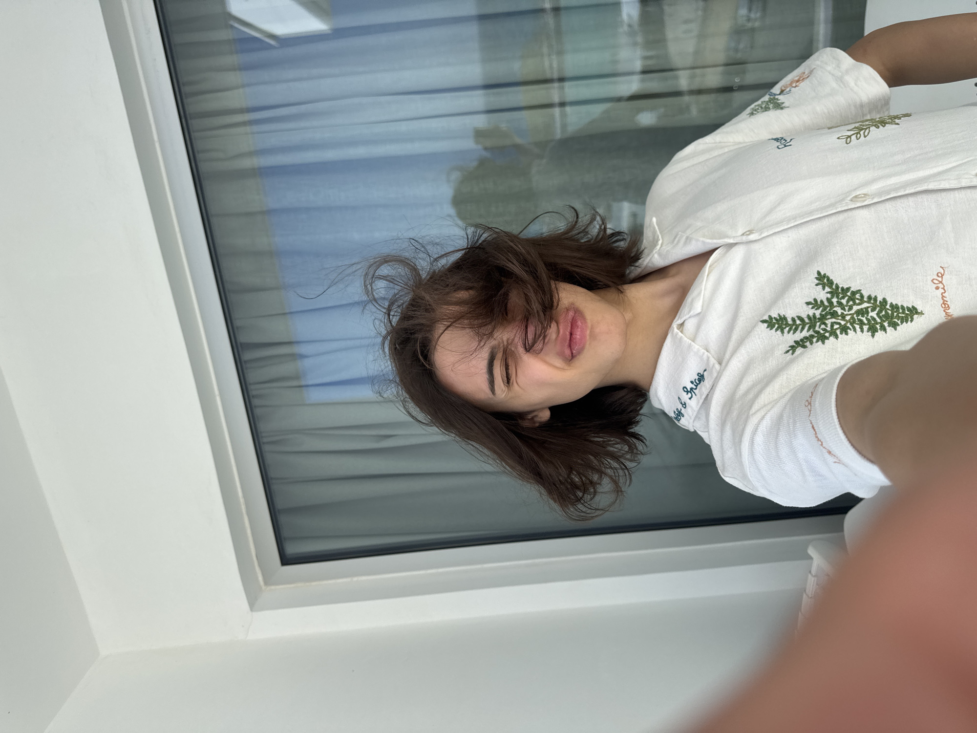 This media file features a close-up, rotated selfie of a young woman taken indoors, likely in Ayia Napa, Cyprus. The image is rotated 90 degrees clockwise, showing the woman's head and upper torso. She has medium-length brown hair, appearing somewhat disheveled and wind-blown, with strands falling across her face. Her eyes are partially closed, and her lips are slightly parted, conveying a relaxed, perhaps playful or sleepy expression.
She is wearing a white collared shirt decorated with green embroidered patterns resembling ferns or trees. Other embroidered elements in brown are also visible. Text is visible on her shirt: "Herb & Spice" is embroidered near the collar/shoulder, and "Chamomile" can be seen next to a brown design on the lower right side. Another partially visible word, possibly "Reel," appears above a green embroidered tree on the right side.
The background consists of a large window with a white frame. The window glass reflects horizontal blinds or curtains, suggesting an interior setting with natural light. The bright lighting indicates it is daytime. A white wall or window sill is also visible. Part of the woman's hand, holding the camera, is seen in the bottom right corner, confirming she is taking a selfie. The overall scene suggests a casual and lighthearted moment.
