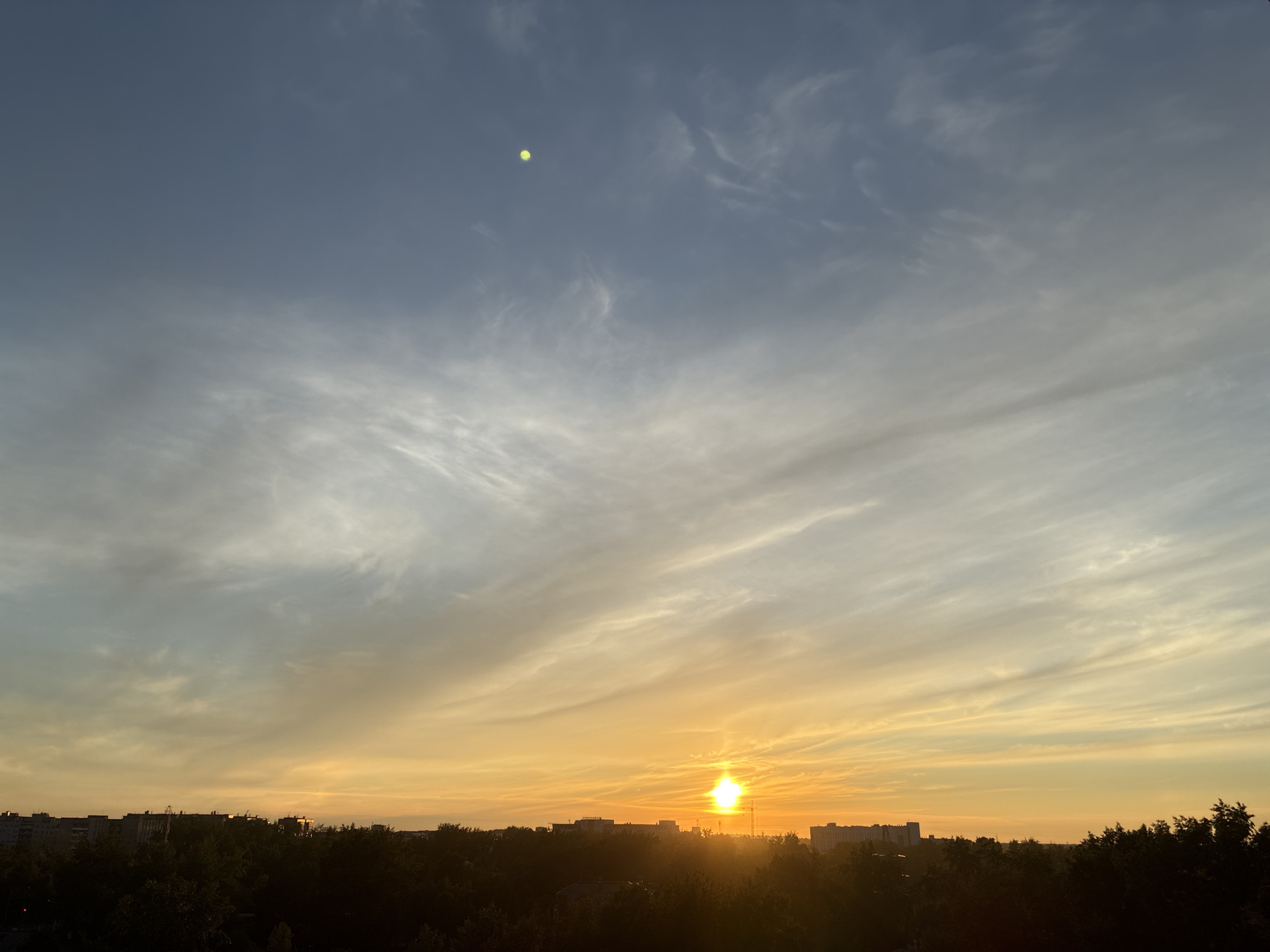 The image captures a sky over a city at sunset. The sky exhibits a gradient of colors, transitioning from a deep blue at the top to shades of orange and yellow near the horizon. Wispy clouds streak across the sky, catching the light of the setting sun. A small, bright yellow object, possibly a balloon or a distant celestial body, is visible in the upper left portion of the sky. Below the sky, a silhouetted treeline runs horizontally across the bottom of the frame, partially obscuring the cityscape. Buildings, appearing as dark shapes against the sky, are visible beyond the trees, suggesting an urban environment. The sun itself is a bright, intense orb low on the horizon, casting its glow over the scene. The overall impression is one of a tranquil evening in an urban setting.