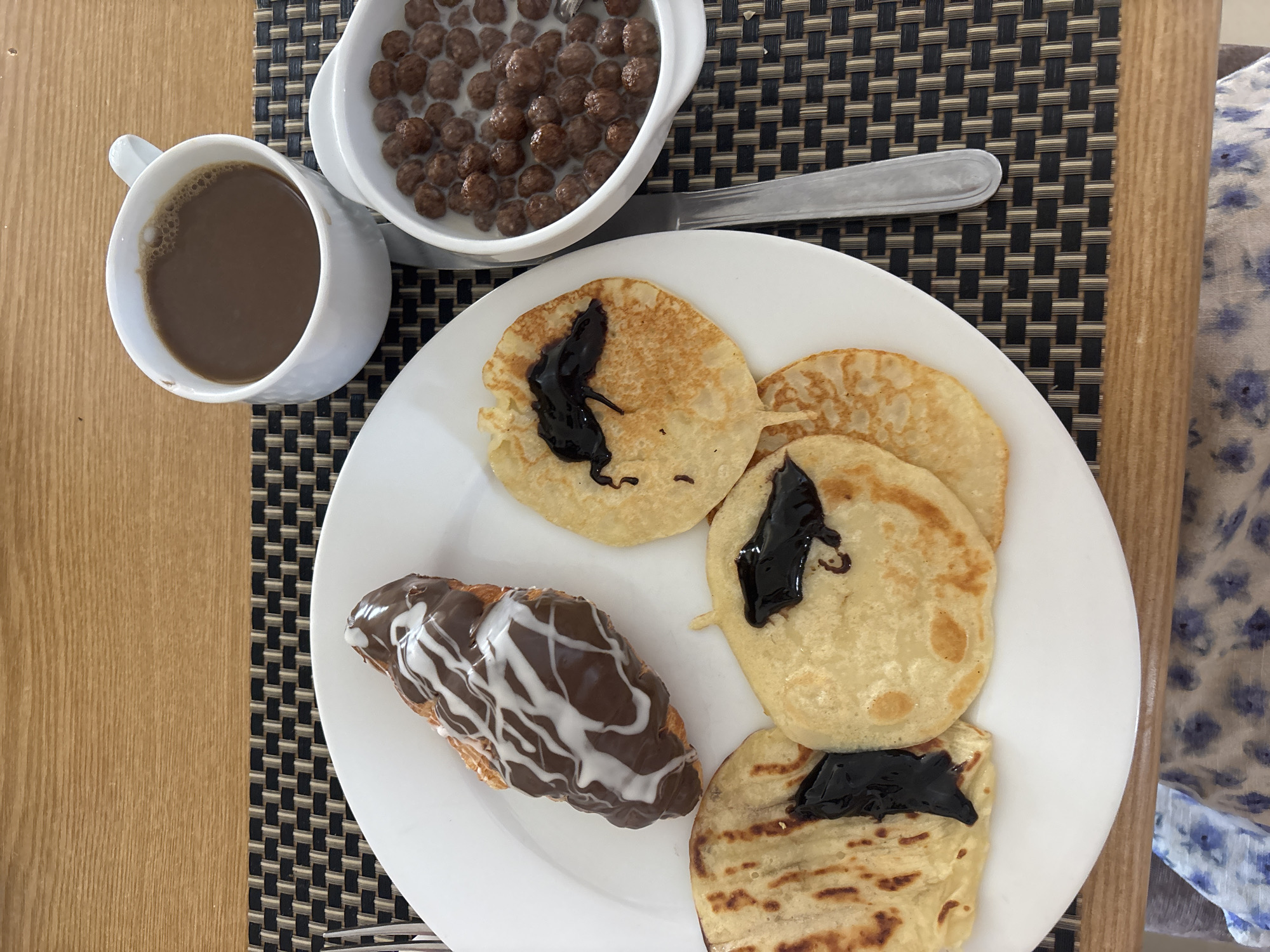 A top-down, close-up shot shows a breakfast meal on a table. The scene includes a white plate with four pancakes, each drizzled with a dark, possibly chocolate, sauce. A chocolate-covered croissant with white icing is also on the plate. A white mug of coffee or hot chocolate sits to the left of the plate. Above the plate is a white bowl filled with chocolate cereal and milk, accompanied by a silver spoon. The table surface has a woven mat with a checkered pattern. The background includes a wooden table and a piece of fabric with a floral pattern. The location is Unknown, Unknown.