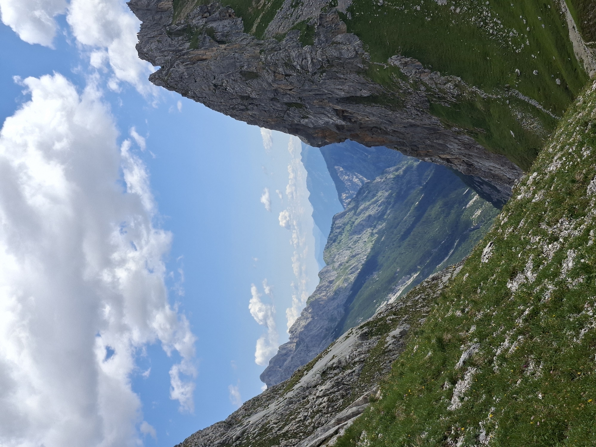 A photograph captures a mountainous landscape with a clear blue sky dotted with white clouds. In the foreground, the rocky slopes of two mountains descend into a valley. The left mountain is covered with green grass and scattered rocks, while the right mountain is predominantly rocky with some patches of green vegetation. A deep valley unfolds between the mountains, revealing a series of more distant, layered mountain ranges that fade into a hazy blue in the background. The overall scene is one of natural, rugged beauty. This image was taken in Wildermieming, Austria.