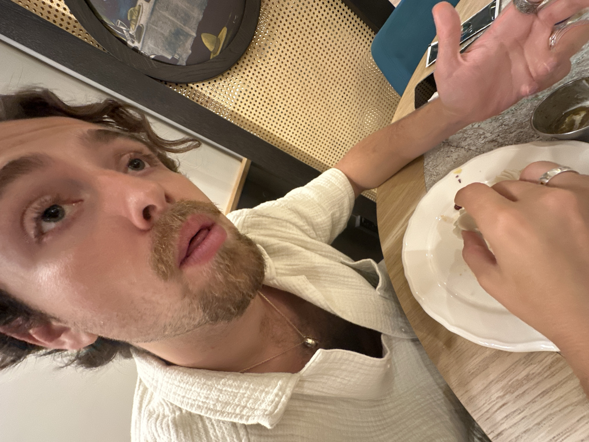 In the city of Belgrade, Serbia, a close-up, high-angle shot captures a person in the foreground wearing a light-colored, short-sleeved shirt. The individual has curly brown hair, a mustache, and a goatee. They are looking directly at the camera, with their mouth slightly open. On their neck, a gold chain with a pendant is visible.
To the right of the person, a black-framed, circular artwork hangs on a wall covered with a woven cane material. The artwork depicts a still life with yellow hats and glasses.
In the lower portion of the image, a white plate with a scalloped edge sits on a wooden table. A hand is reaching into the plate, holding what appears to be food. The hand is adorned with two silver rings. To the right of the plate, another hand is open, palm facing upwards.
A small metal cup containing a greenish liquid is visible on the table, as well as a black object, possibly a phone, and a white packet. The background shows a glimpse of a teal-colored chair. The overall scene suggests a dining setting.