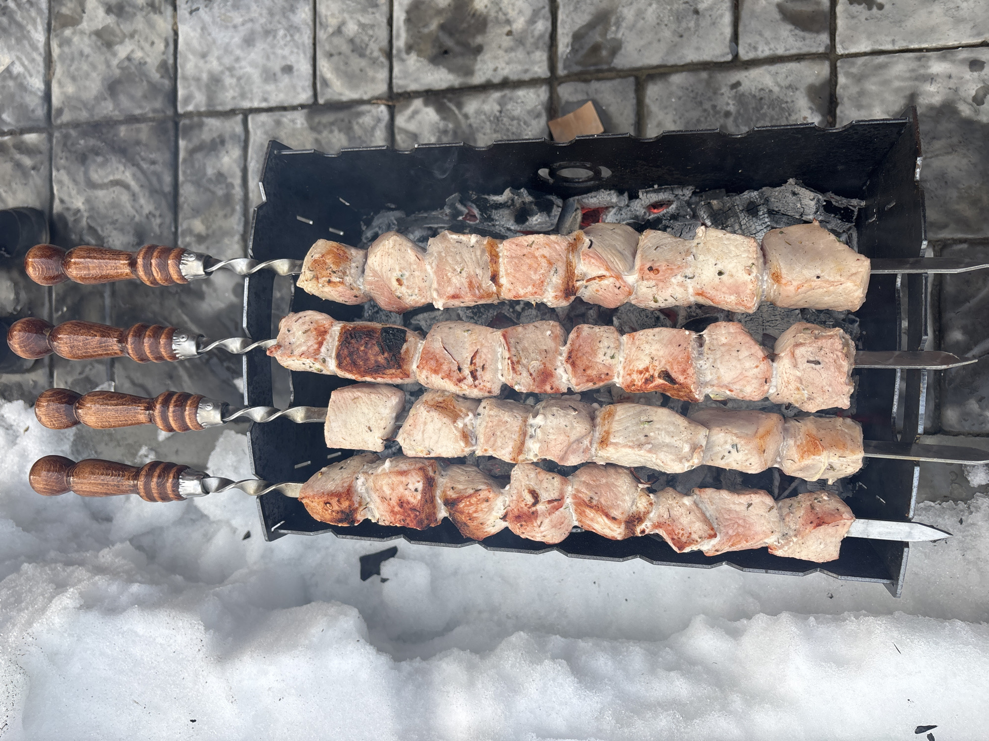 Four metal skewers with cubed meat cook over glowing coals in a black metal brazier. The meat exhibits varying degrees of doneness, from light pink to browned and lightly charred, with visible seasoning. Each skewer features a twisted metal shaft and a dark, carved wooden handle. The brazier is positioned outdoors in Chaikovsky, Russia. To the left, a large, undisturbed mass of white snow is present. To the right, a surface composed of wet, grey rectangular paving stones is visible. Smoke rises from the brazier.
