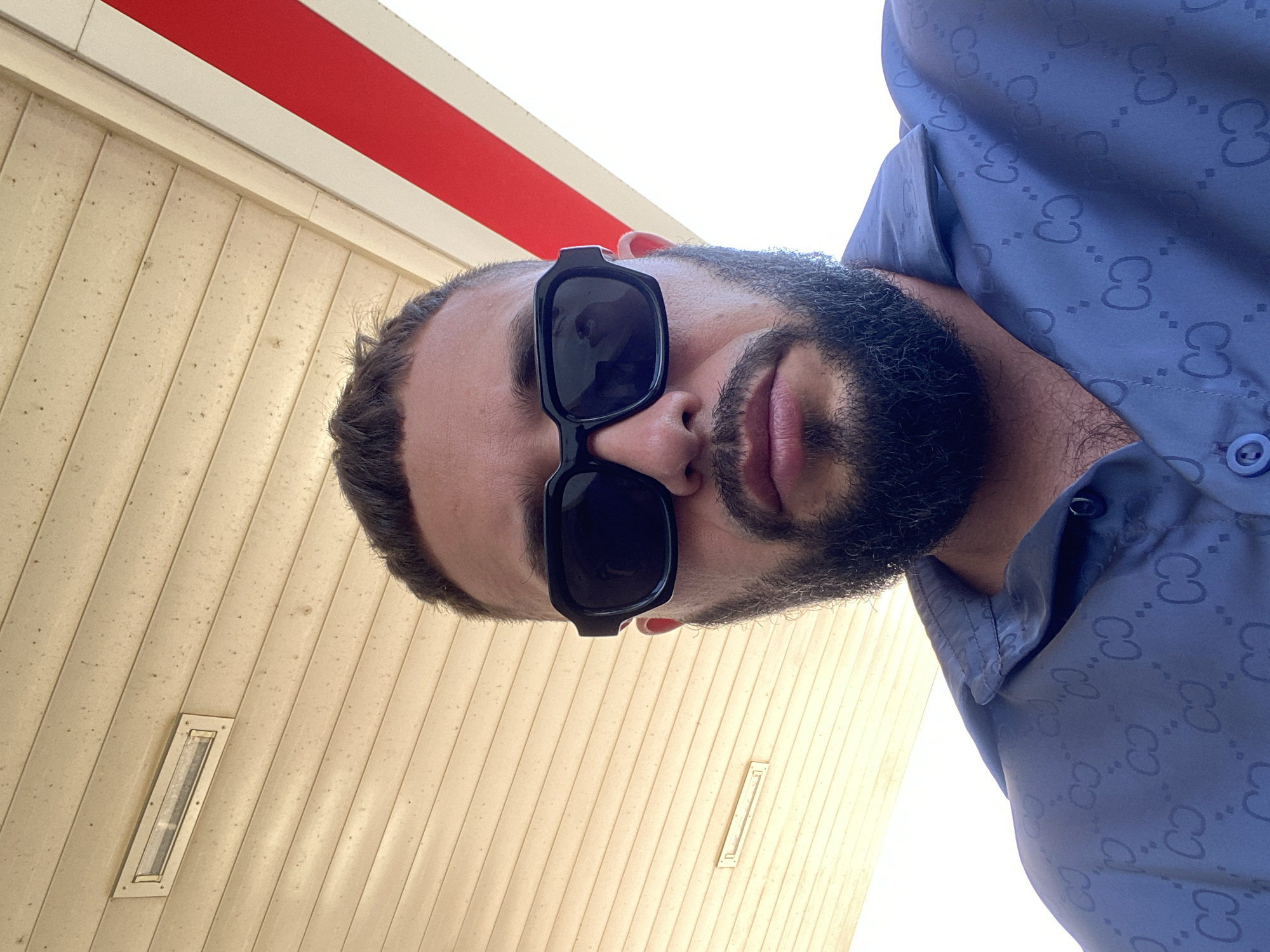 A man wearing black sunglasses and a blue patterned button-up shirt is positioned in the center of the frame, looking upwards towards the camera. The background consists of a textured ceiling and a white and red stripe. A rectangular light fixture is visible on the ceiling. The image appears to have been taken outdoors in a location with overhead cover, possibly at a gas station or a covered walkway. The man has a beard and short hair. The overall scene is a close-up portrait against an architectural backdrop.