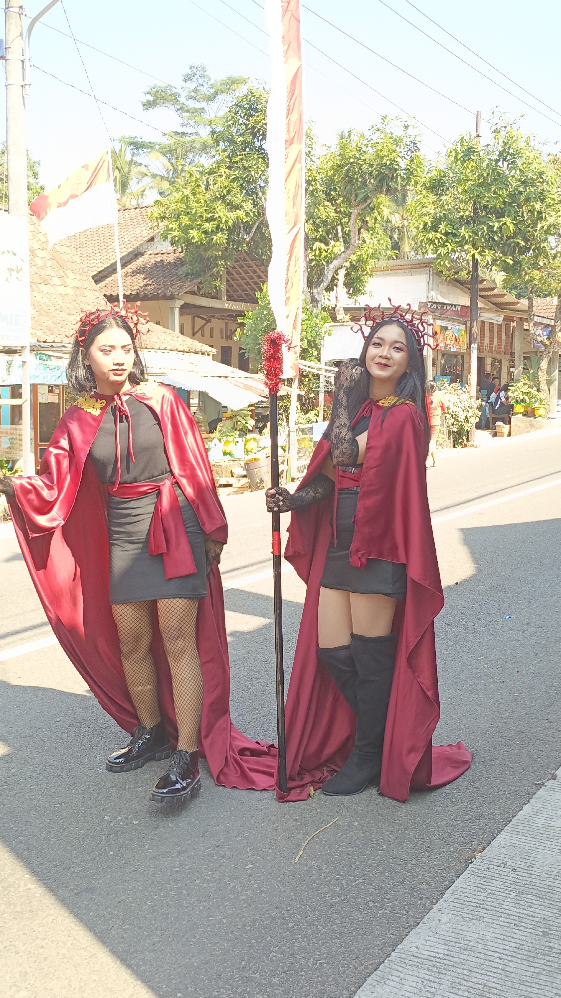 Two women are posing for a photo on a paved road in Krajan Lor, Indonesia. The woman on the left is wearing a black dress, fishnet stockings, and black platform boots, with a long red cape draped over her shoulders. She has a red crown-like headpiece and her hair is pulled back. The woman on the right is wearing a black skirt, black thigh-high boots, and a black lace top with a red cape over her shoulders. She also has a red crown-like headpiece and her hair is down. She is holding a staff with a red, spiky top. Both women are looking towards the camera. The background shows a street with buildings, trees, and power lines. An Indonesian flag is visible on the left side of the image. The sun is shining, casting shadows on the road.