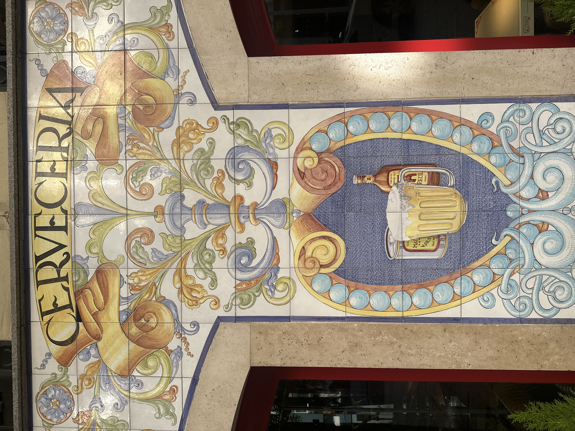Tile artwork of a storefront in Madrid, Spain. The word "CERVECERIA" is vertically displayed in black capital letters, with a golden shadow effect, on a tile artwork. The artwork depicts a beer bottle and mug within an oval frame, adorned with blue orbs. Stylized scrolls and floral ornamentation are also part of the design, all created with glazed ceramic tiles. The artwork is set above a window frame.