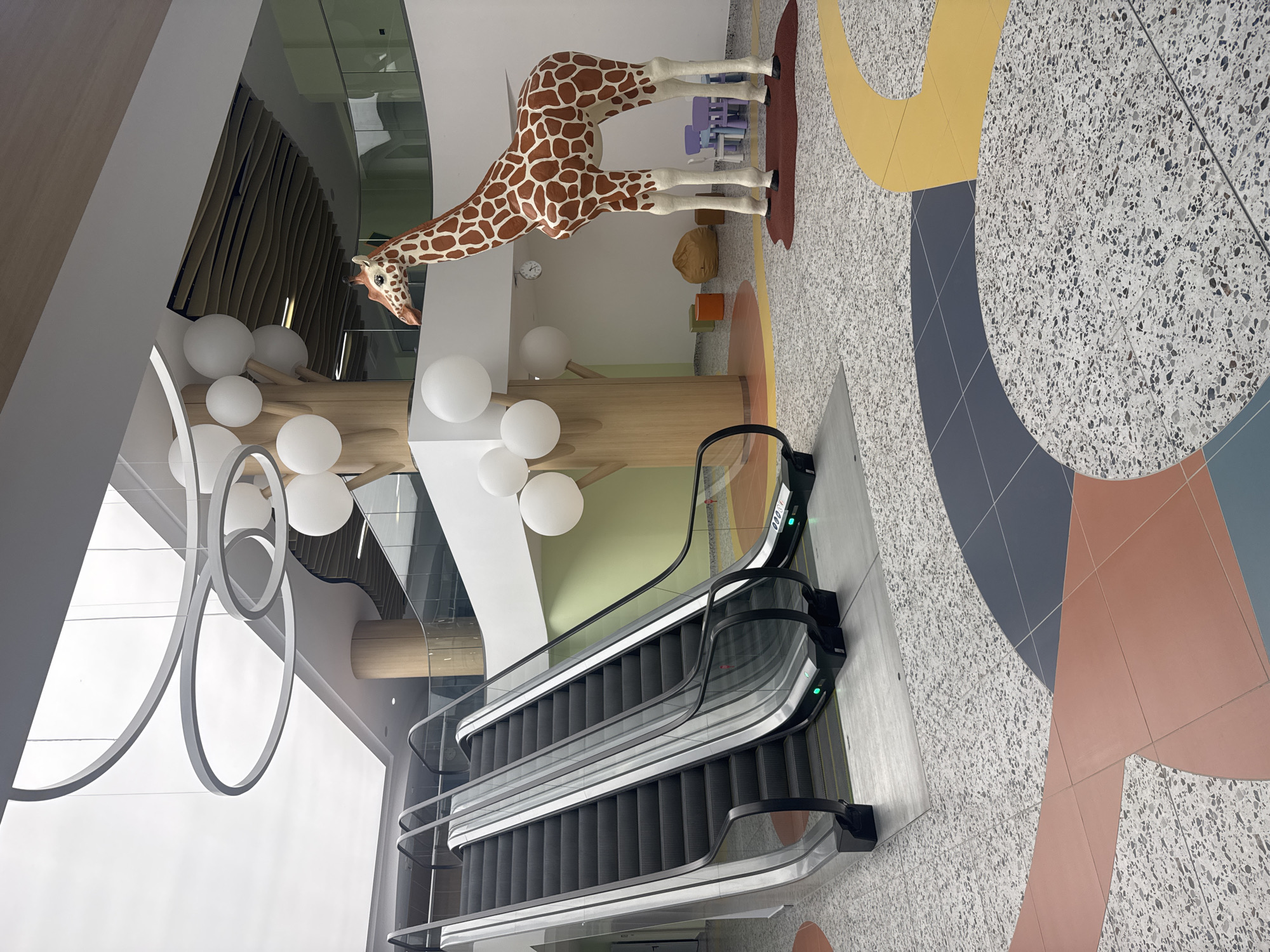 The image depicts the interior of a modern, brightly lit building, likely a public space or a children's facility, given the presence of a large giraffe statue and playful decor. Two escalators are prominently positioned on the left side of the frame, both angled upwards and appearing to be in operation with green indicator lights visible at their bases. The floor is a multi-colored terrazzo with large, abstract shapes in shades of gray, orange, and yellow.
To the right of the escalators, a life-sized statue of a giraffe stands, its distinctive orange and brown spotted pattern vividly rendered. Behind the giraffe, there is a children's play area with small, colorful chairs and a beanbag chair. A wooden pillar with tree-like branching structures, adorned with white spherical decorations resembling large balloons or clouds, is situated behind the giraffe. Suspended from the ceiling are modern, circular pendant light fixtures. The walls are painted in soft pastel colors, including a pale green in the background. The overall atmosphere is clean, modern, and inviting, with a focus on playful design elements.
This scene is located in the new wing of the Burgas Central Hospital, Burgas, Bulgaria.