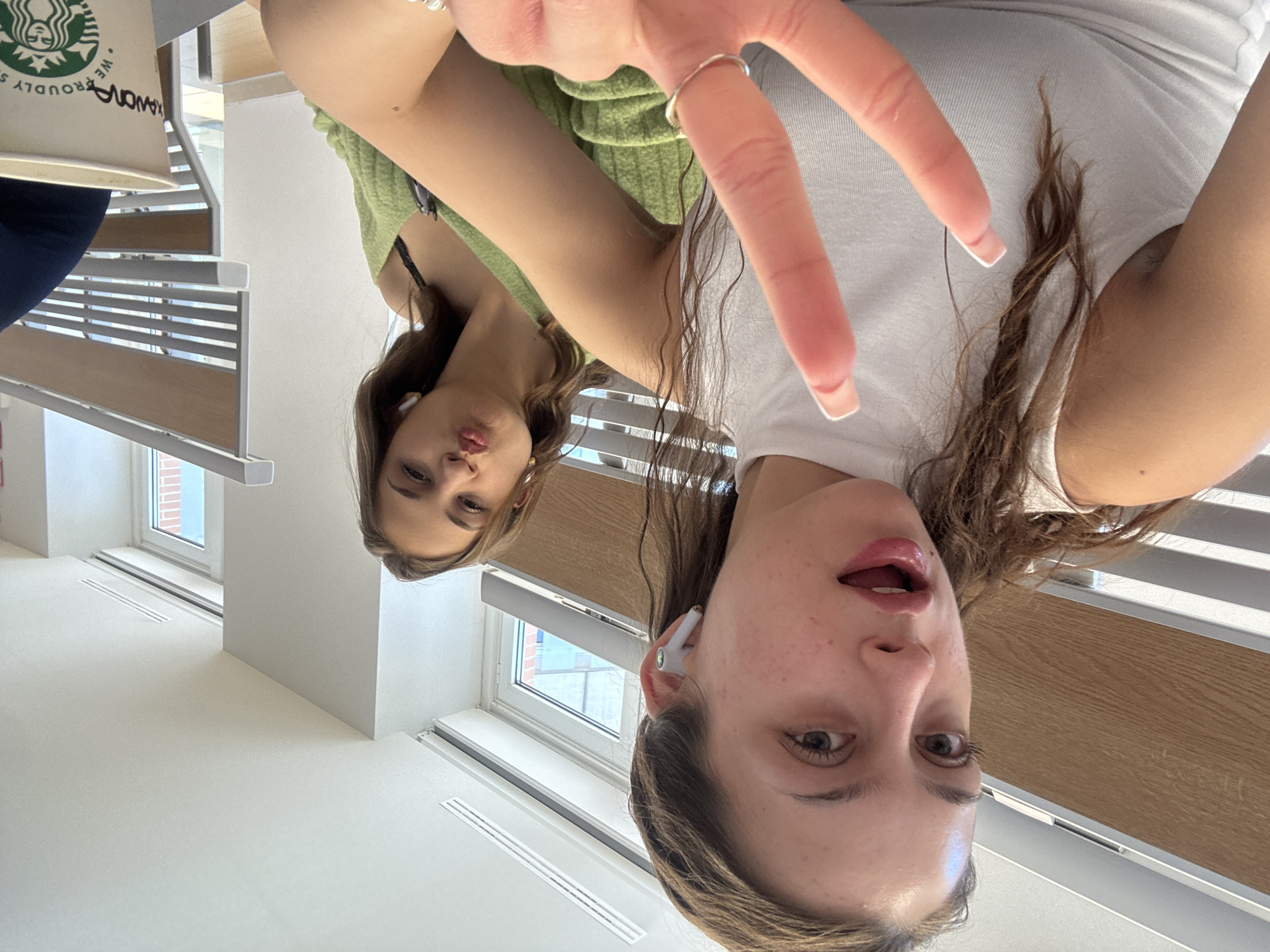 Two women are posing indoors. One is wearing a light green top, and the other is wearing a white t-shirt. The woman in the white shirt is making a peace sign with her hand. There is a Starbucks cup visible in the background. The indoor location is indicated by windows, walls, and wooden slats that appear to be part of a window blind or a bench. Based on the style and design of the interior, and the presence of the Starbucks cup, it is likely this photo was taken at the Starbucks Reserve Roastery in Milan, Italy.