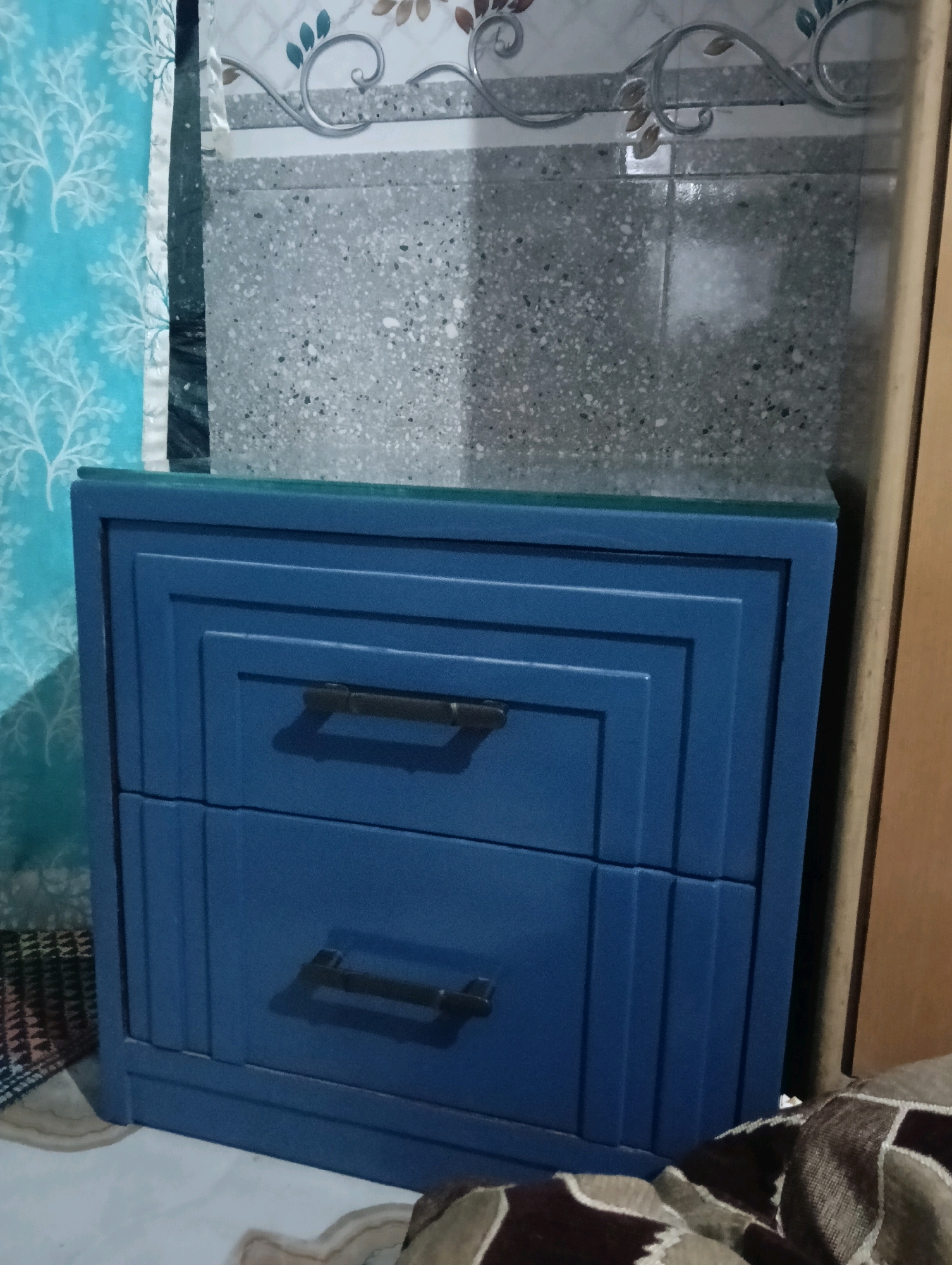 A blue, two-drawer nightstand with rectangular metal handles is positioned in a room. The nightstand has a layered, recessed front design. A patterned blue curtain hangs to the left of the nightstand. Behind the nightstand, a wall is visible, decorated with tiles featuring a speckled grey pattern and an ornate floral and scroll design in white and brown. The flooring appears to be a light-colored tile or linoleum. The scene suggests a domestic interior. There is no indication of the specific location within the city of Mumbai, India.