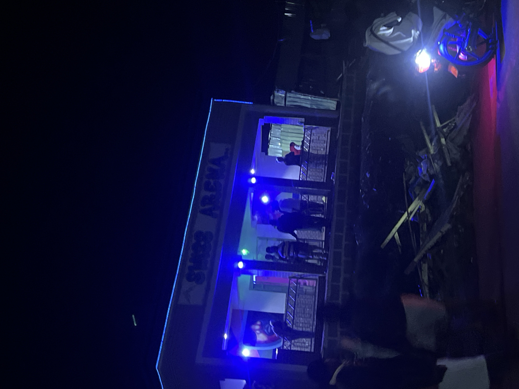 The image depicts a nighttime scene in Durumi, Nigeria, featuring a building with "SWISS ARENA" signage illuminated by blue neon lights. Several people are visible on a raised walkway in front of the building, appearing to be gathered or socializing. The area in front of the building seems to be a street or open space, with debris and discarded materials scattered on the ground, suggesting potential construction or recent damage. To the right of the frame, a motorcycle is parked, its headlights casting a bright glow and reflecting pink and blue light on the ground. The overall ambiance is one of a bustling nightlife venue situated in a somewhat unkempt urban environment.