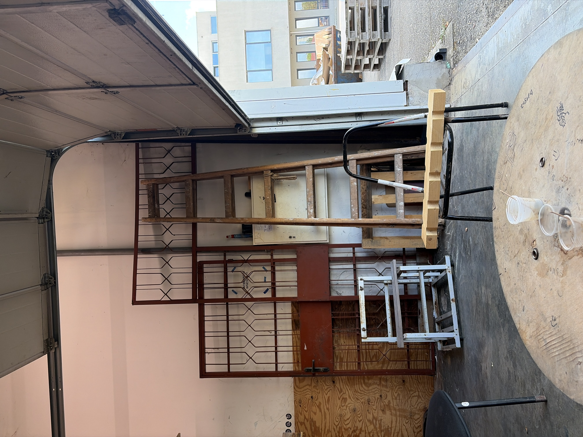 This image depicts an interior scene within a large garage or workshop space, likely located in Ravno pole, Bulgaria. The space appears to be partially open to an exterior area, indicated by the large overhead garage door which is raised. Inside, various items are stored, suggesting a functional or storage area. Prominently featured is a tall wooden ladder leaning against the back wall. To the left of the ladder are two large, red metal gates or doors with decorative lattice work. In front of these, a small, white metal shelf unit on wheels is visible. A wooden table, possibly a spool from a cable roll, occupies the foreground, with two plastic cups and a ring on its surface. Another black chair with a wooden element rests in front of the ladder. In the background, outside the garage, a modern building with large windows and a dumpster filled with debris can be seen, along with stacked wooden pallets. The floor is concrete. The overall impression is of a cluttered but organized storage or work area.
