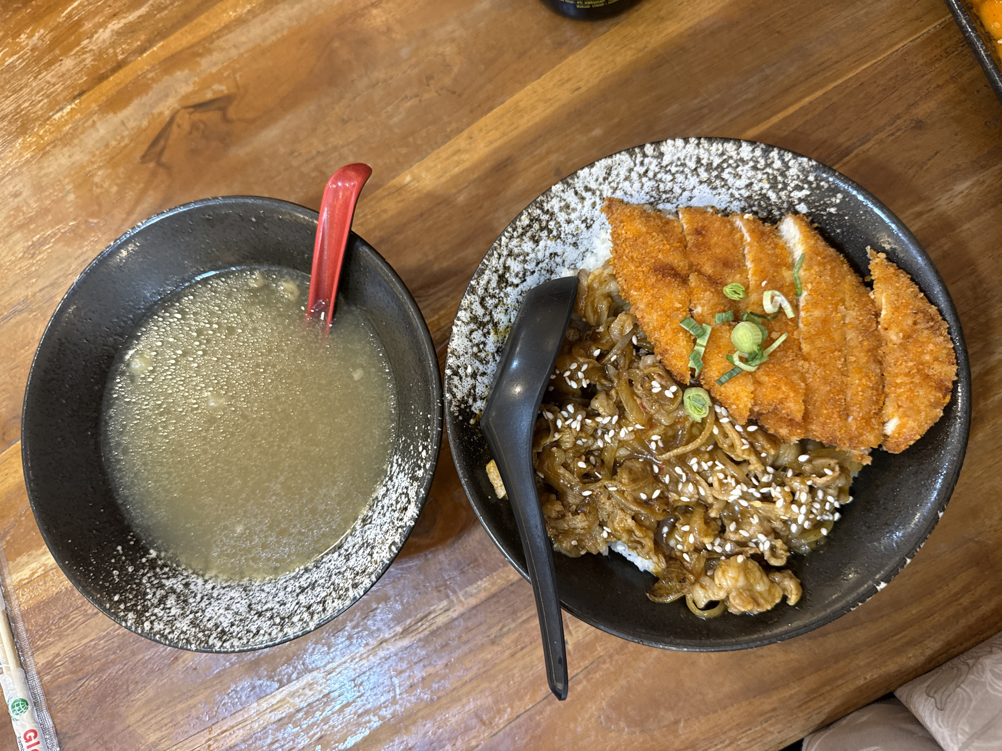 The image shows a top-down view of a wooden table setting with two bowls of food. The bowl on the left contains soup, and there is a red spoon inside. The bowl on the right holds rice topped with beef, sesame seeds, and cutlets, likely chicken katsu, garnished with sliced scallions. A black spoon is placed in the bowl on the right. Chopsticks are visible on the left side of the image. The bowls are black with a speckled design around the edges. Considering the food style, it is likely that the location of the image is a restaurant or dining establishment. There are no people visible. The image does not indicate a specific location within Lhokseumawe, Indonesia.