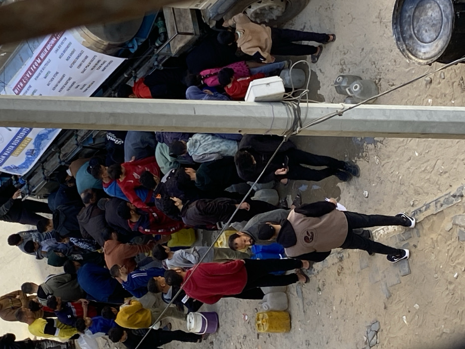 An aerial view depicts a crowded water distribution scene in Khan Yunis, Palestinian Territory. Numerous individuals, predominantly men and children, are gathered around a stationary water tanker truck. The truck's cylindrical tank has two white banners attached; one reads "CLEAN WATER DISTRIBUTION" and "YOUTH DAY," while the other, partially obscured, displays "WITH LOVE FROM MILLIE BROWN" and a list of names. People are actively collecting water into various containers, including yellow, blue, and clear plastic jerrycans, white buckets, and smaller bottles, many bending over near the truck's lower outlets. The ground is unpaved, appearing as compacted dirt or sand with visible wet patches and some debris. In the foreground, a utility pole with multiple wires runs vertically. A man in a tan vest with a dark logo is visible from behind, holding papers. Another bearded man stands near blue containers, observing the distribution.