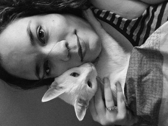 A black and white, grainy close-up image depicts an adult female subject holding a white cat. The female, positioned on the right side of the frame, has dark, wavy hair and looks towards the viewer with a slight, closed-mouth smile. A thin strand of hair crosses her nose. Her left hand, visible in the lower left quadrant, displays a ring with multiple small stones on the ring finger. She wears a striped garment, partially visible on her right side. The white cat, possessing dark eyes and prominent ears, is nestled against the woman's left cheek, its head turned away from the camera. Its body is partially obscured by the woman's arm and a patterned fabric, possibly a blanket or bedspread, visible below. The background is an undifferentiated dark surface, indicative of an indoor setting. The image provides no discernible visual information to identify a specific location within Buenos Aires, Argentina.