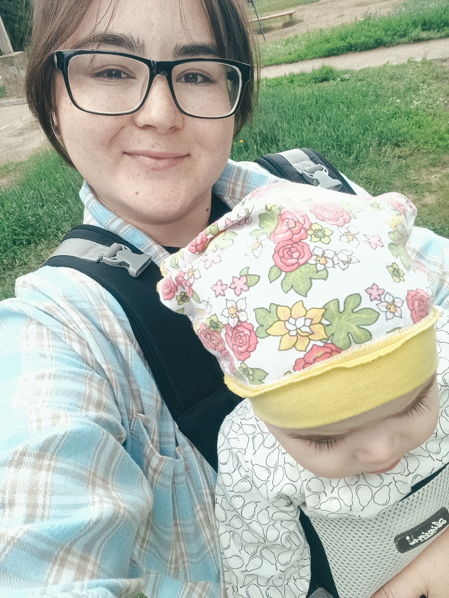 A person with glasses is taking a selfie while holding a baby in a carrier. The person is wearing a blue and white plaid shirt. The baby is wearing a floral print hat and a white onesie with a pear pattern. The background appears to be a grassy outdoor area, possibly a park or yard, with a dirt path and a bench visible. The overall scene suggests a casual outing. The specific location within the city of Noviy, Russia, cannot be determined from the visual information alone.