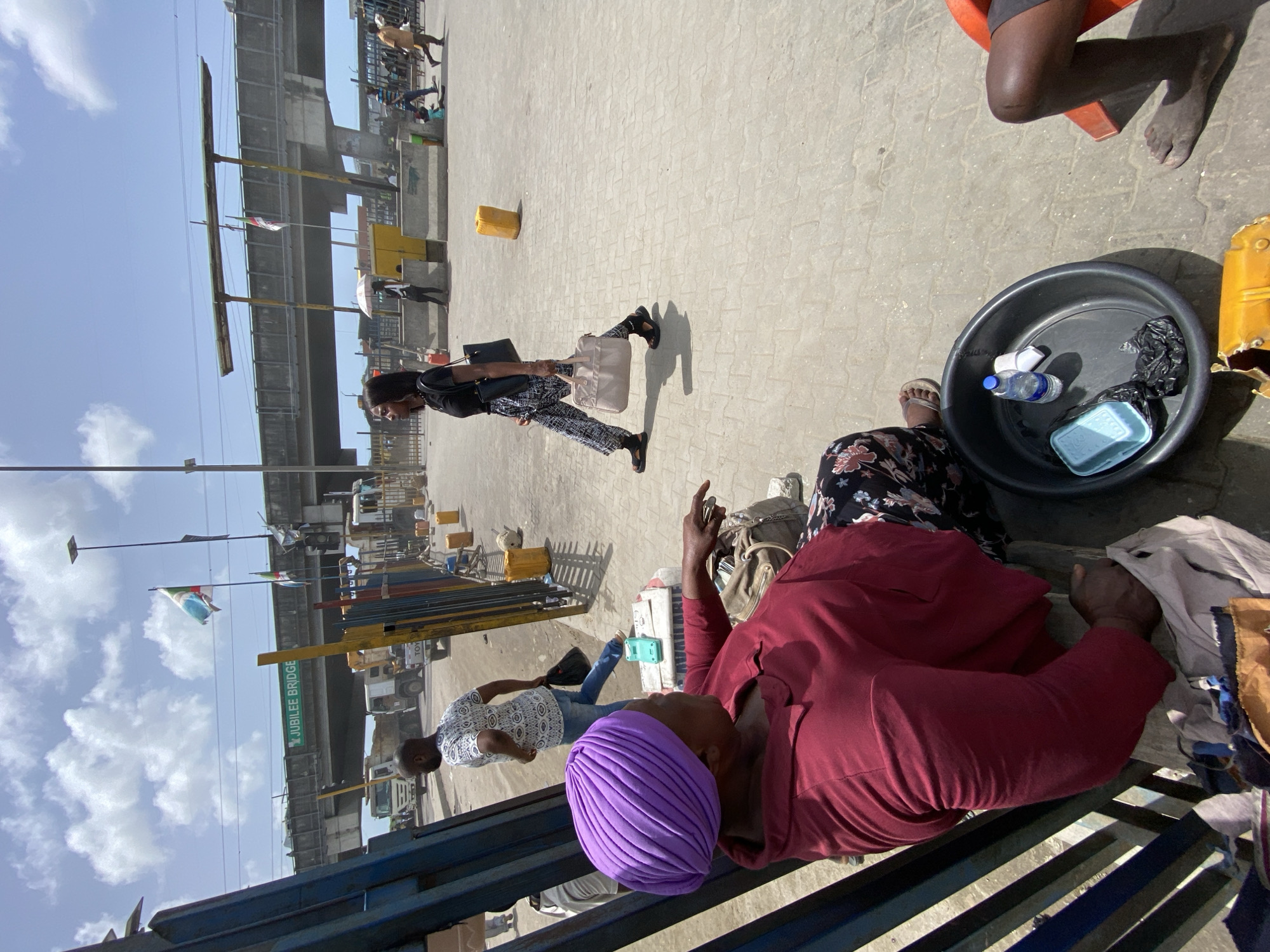 The image captures a street scene near the Jubilee Bridge in Lekki, Nigeria. The environment is urban, with an elevated bridge structure visible in the background. A woman wearing a purple headwrap and a maroon shirt is seated on a blue metal structure, holding clothing in her hands. Nearby, a black tub contains a water bottle and other items. Another woman wearing a patterned outfit sits next to the tub. A woman dressed in black and patterned pants is walking away from the camera with a bag. Additional individuals are visible in the background.