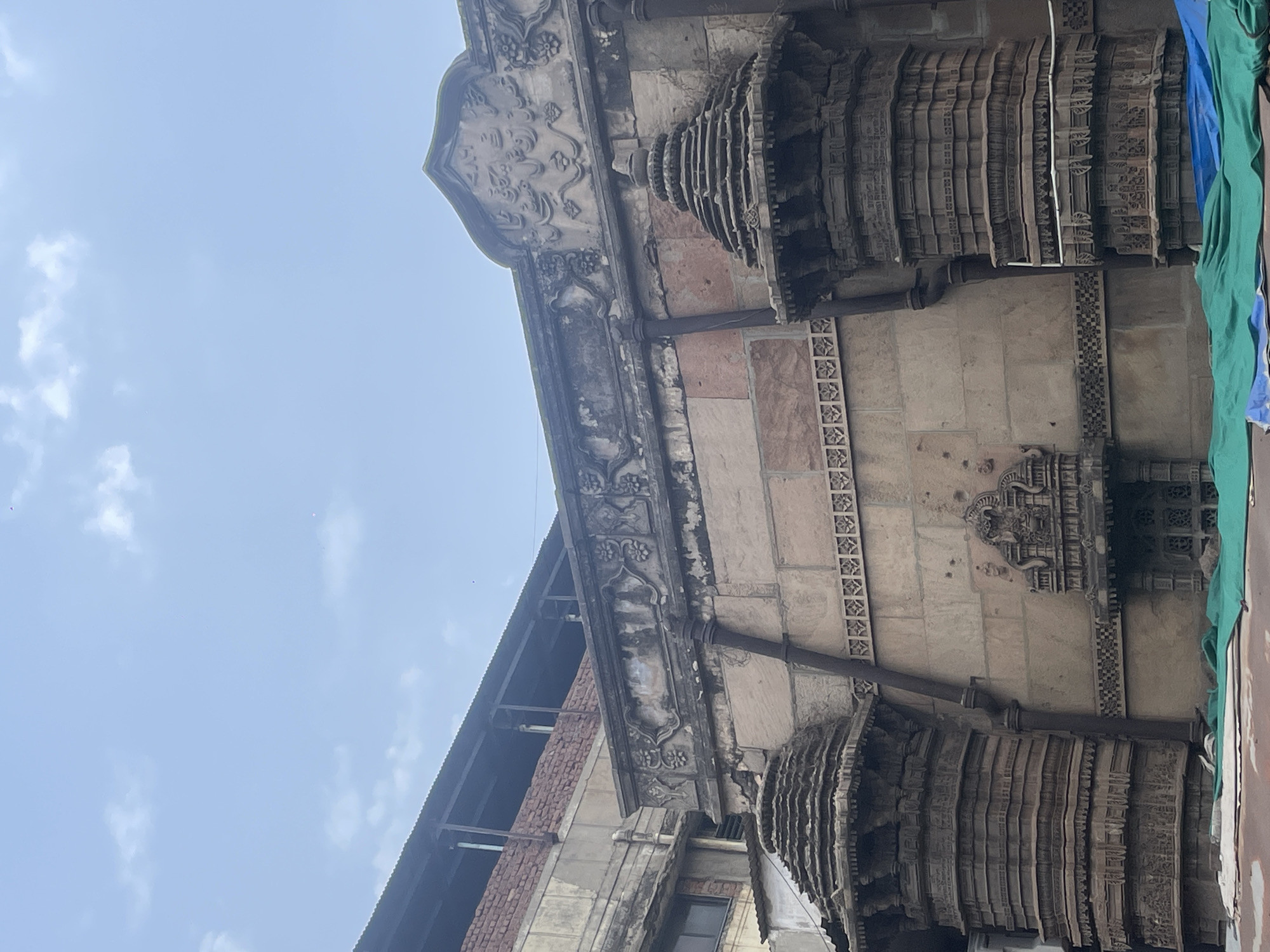 This is a vertical, eye-level, outdoor shot of an ancient stone building in Ahmedabad, India. The building features intricate carvings and architectural details, including ornate pillars and decorative friezes. The stone appears weathered and has a reddish-brown hue in some sections, contrasting with areas of grey stone. A dark, possibly rusted, pipe runs vertically down the facade. In the foreground, a bright green tarp is draped over some unseen objects, suggesting ongoing work or protection. The sky above is a pale blue with scattered white clouds. The architectural style points to a historical structure, likely a temple or mosque, given the region.