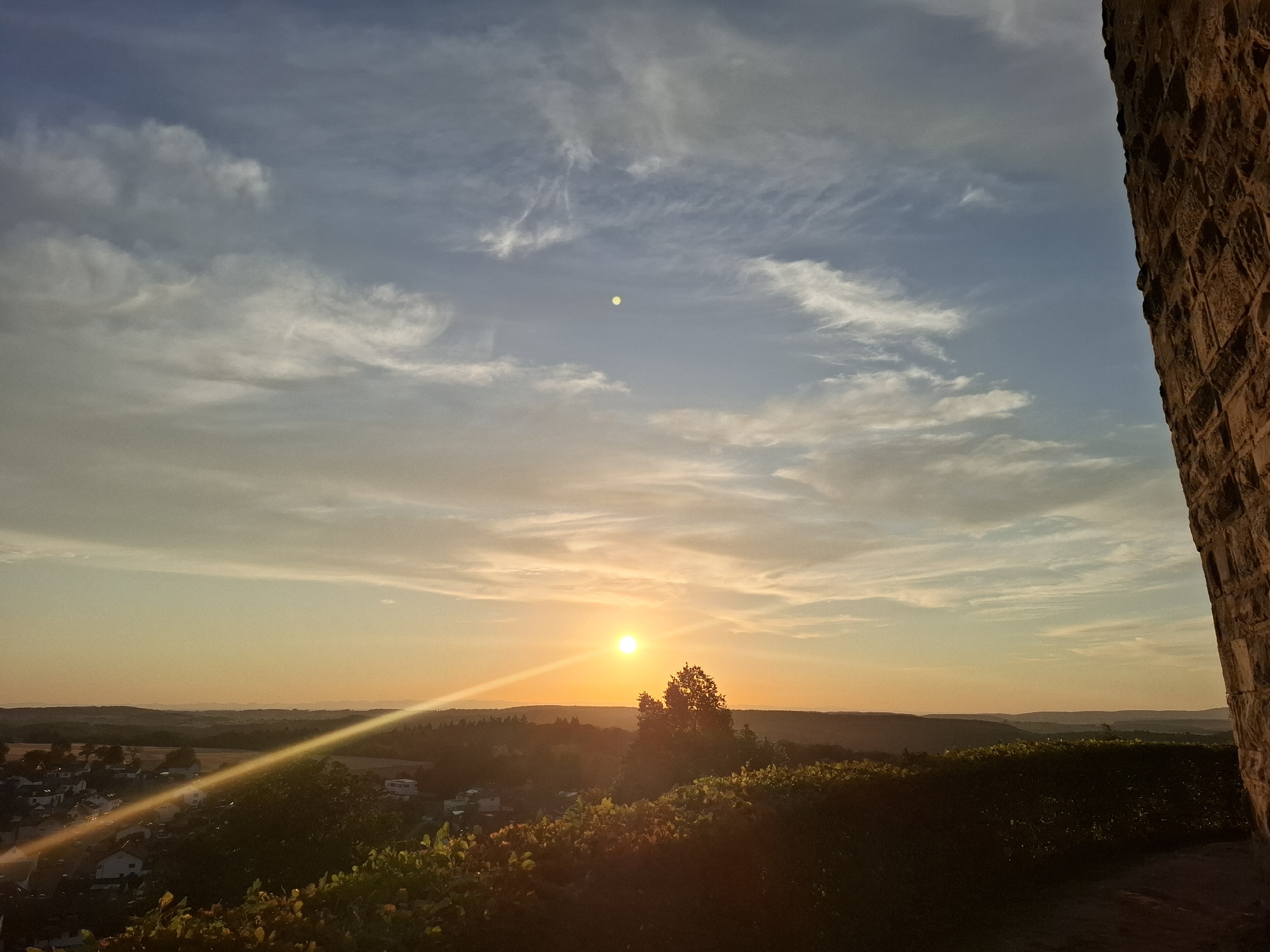 This image depicts a sunset viewed from the grounds of the Sinsheim Castle ruins in Sinsheim, Germany. The sun is low on the horizon, casting a warm, golden light across the sky and the landscape below. Wispy clouds, tinged with orange and pink, stretch across a blue sky. A faint, pale yellow disc, likely the moon or a bright planet, is visible in the upper portion of the sky.
In the foreground, the rough, textured stone of the castle wall is visible on the right side of the frame, occupying about a quarter of the image. Below and to the left of the castle wall, dense green foliage, possibly a vineyard or garden, is silhouetted against the sunset.
The middle ground reveals a rolling landscape with scattered trees and the roofs of buildings belonging to a small town or village. A prominent, bright sunbeam cuts diagonally across the scene from the lower left to the upper right, creating a lens flare effect. In the distance, layers of hills fade into the hazy horizon. The overall scene is one of natural beauty and historical ambiance. There are no people or animals visible in the image.