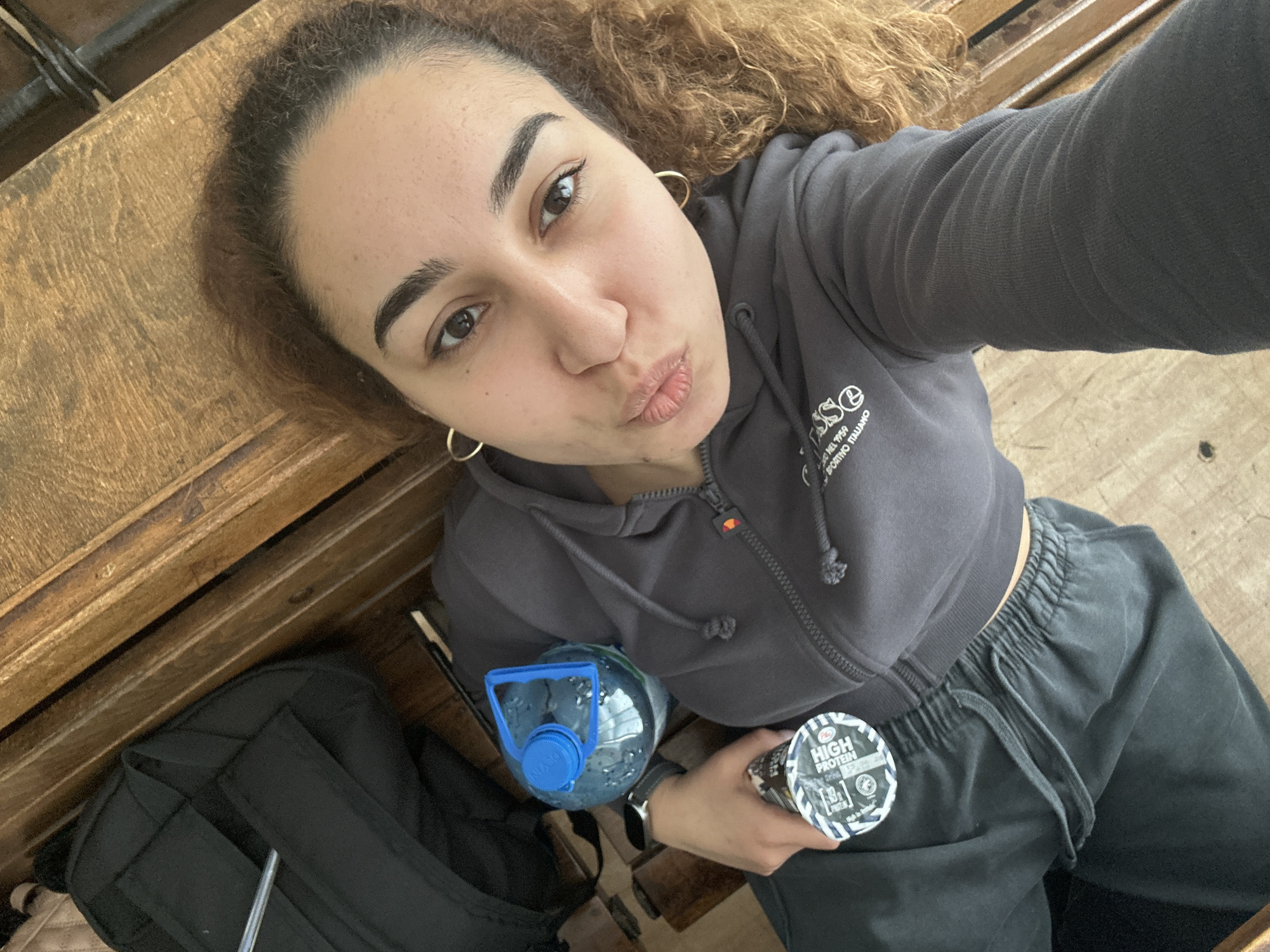A young woman is taking a selfie while lying on a wooden surface. She is fair-skinned with brown curly hair and is wearing a gray hoodie and black sweatpants. She has hoop earrings in both ears. In her right hand, she is holding a high-protein drink container, and to her left is a blue water bottle. A black backpack is partially visible next to her. The location is unknown, and there is no indication of Burgas, Bulgaria.