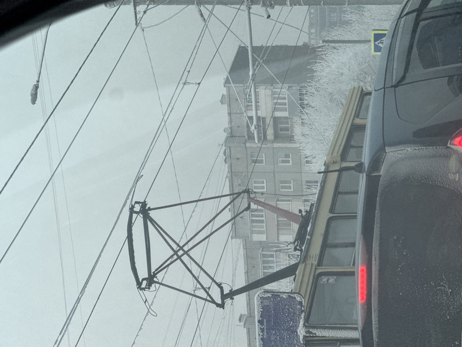 The scene is an urban winter environment in Krasnoyarsk, Russia. A dark-colored vehicle occupies the lower foreground, presenting its snow-dusted rear hatch and illuminated red brake lights. A black whip antenna extends from its roof. Immediately behind, the upper portion of a light-colored (yellow/cream) public transit vehicle, identified as a trolleybus or tram, is visible. Its pantograph, a dark metallic structure with red supporting components, extends vertically to contact overhead electrical lines, which are themselves coated in frost. A blue sign, partially obscured by snow, is mounted on the side of the transit vehicle. Dense frost covers leafless trees and utility poles visible throughout the midground and right frame. A frosted streetlamp fixture is present in the upper right. In the background, multiple grey, multi-story residential buildings with rectangular windows and balconies are discernible under a uniformly overcast, light grey sky. No human figures are present.