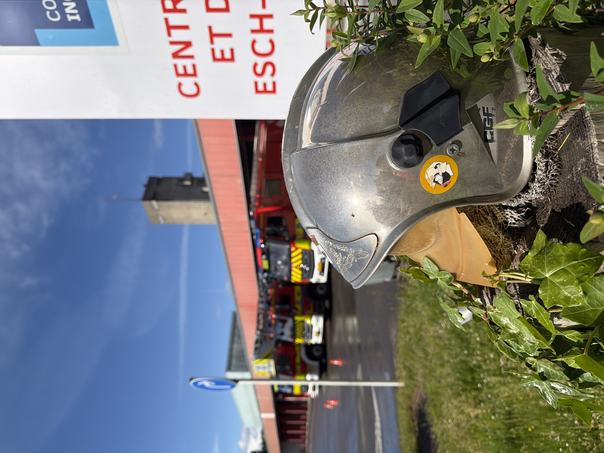 The image shows a fire helmet placed on a wooden post covered in ivy, located in front of the Centre d'Incendie et de Secours Esch-sur-Alzette fire station in Luxembourg. Several fire trucks are parked in the background. A sign is visible to the left. The sky is blue with a few clouds.