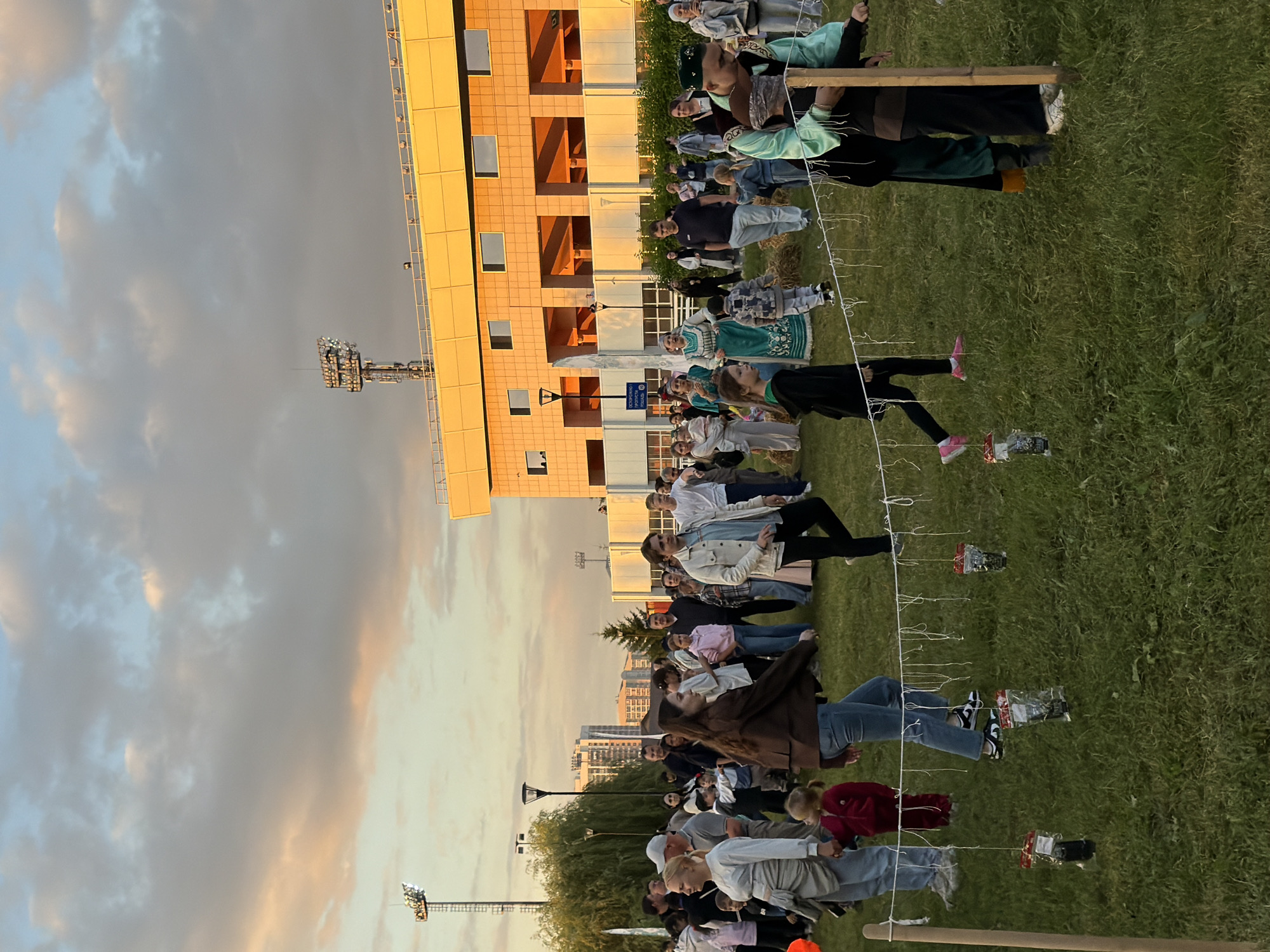 A group of people are gathered on a grassy field in front of a modern building with orange accents, possibly a stadium or arena. The sky is filled with dramatic clouds, illuminated by the warm glow of sunset. In the foreground, a rope is strung between two wooden posts, with small bags hanging from it. Several people are walking around the field, some in casual attire, while others are wearing more traditional clothing, suggesting a cultural event. Children are also present, some holding hands with adults. The overall scene depicts a lively outdoor gathering during twilight.