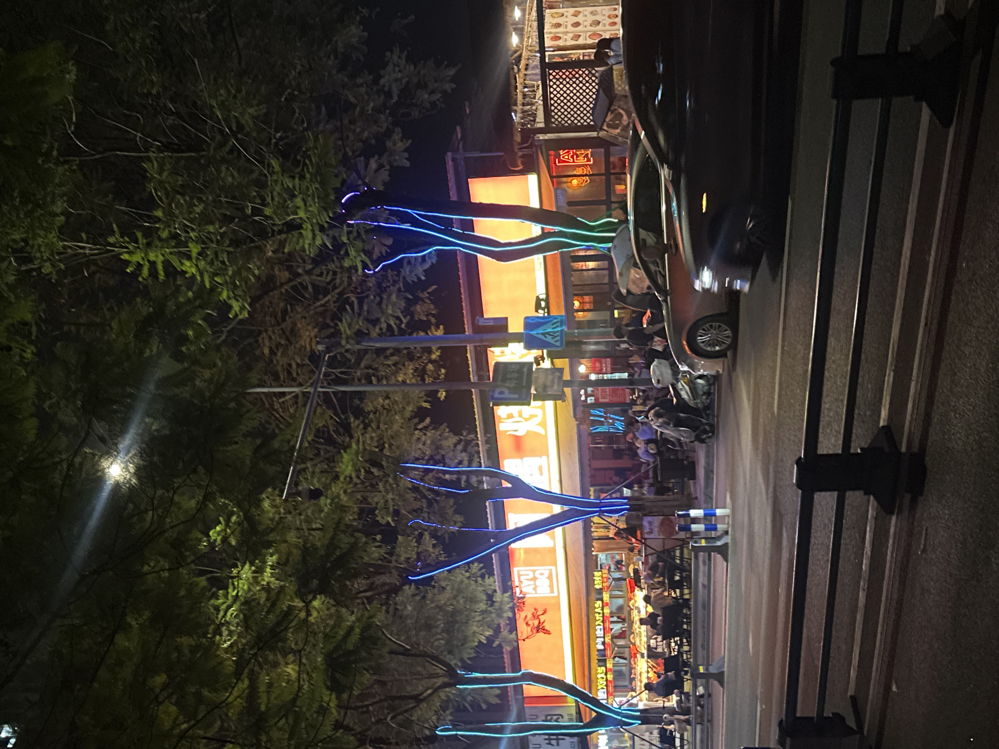 The image depicts a street scene at night in the Chongqing High-tech Zone, China. A row of restaurants or food stalls lines the street, illuminated by bright orange and red neon signs. Trees with blue neon lights wrapped around their trunks and branches stand in front of the establishments. Several people are seated at tables outside the eateries, engaged in dining and conversation. Cars are parked and driving on the street in the foreground. The overall atmosphere is lively and typical of a commercial district at night.