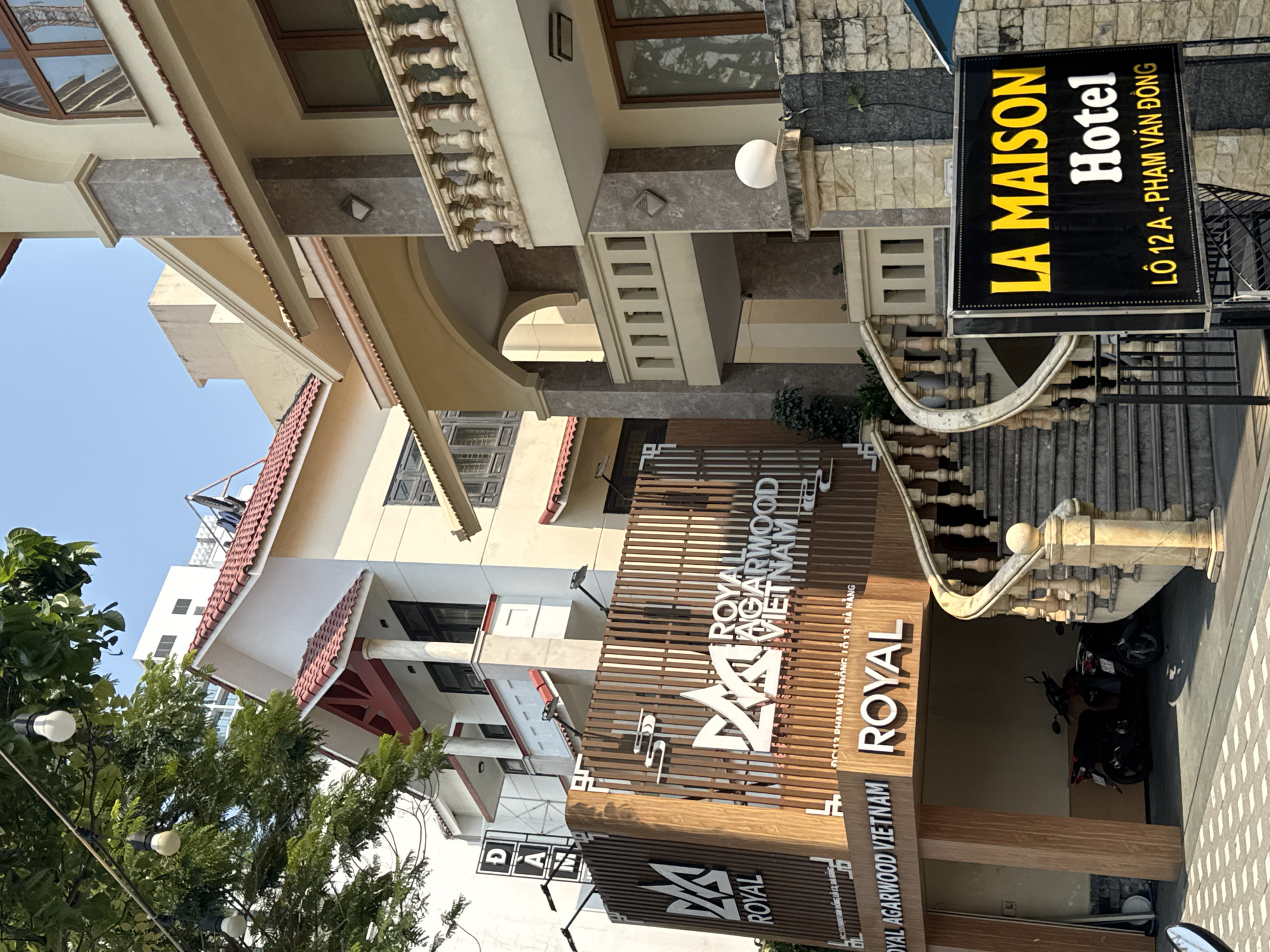 The image depicts the exterior of two establishments in Đà Nẵng, Vietnam: Royal Agarwood Vietnam and La Maison Hotel.
Royal Agarwood Vietnam features a brown wooden structure with a large "VA Royal" logo. The sign indicates the address as 12 Phạm Văn Đồng, Lô 13, Đà Nẵng.
La Maison Hotel has a yellow sign with black lettering and lists the address as Lô 12 A - Phạm Văn Đồng. A curved, ornate staircase with a stone balustrade leads up to the hotel. Several parked motorbikes are visible under the staircase.
The surrounding buildings are multiple stories tall, and partially obscured by trees. The sky is a clear blue.