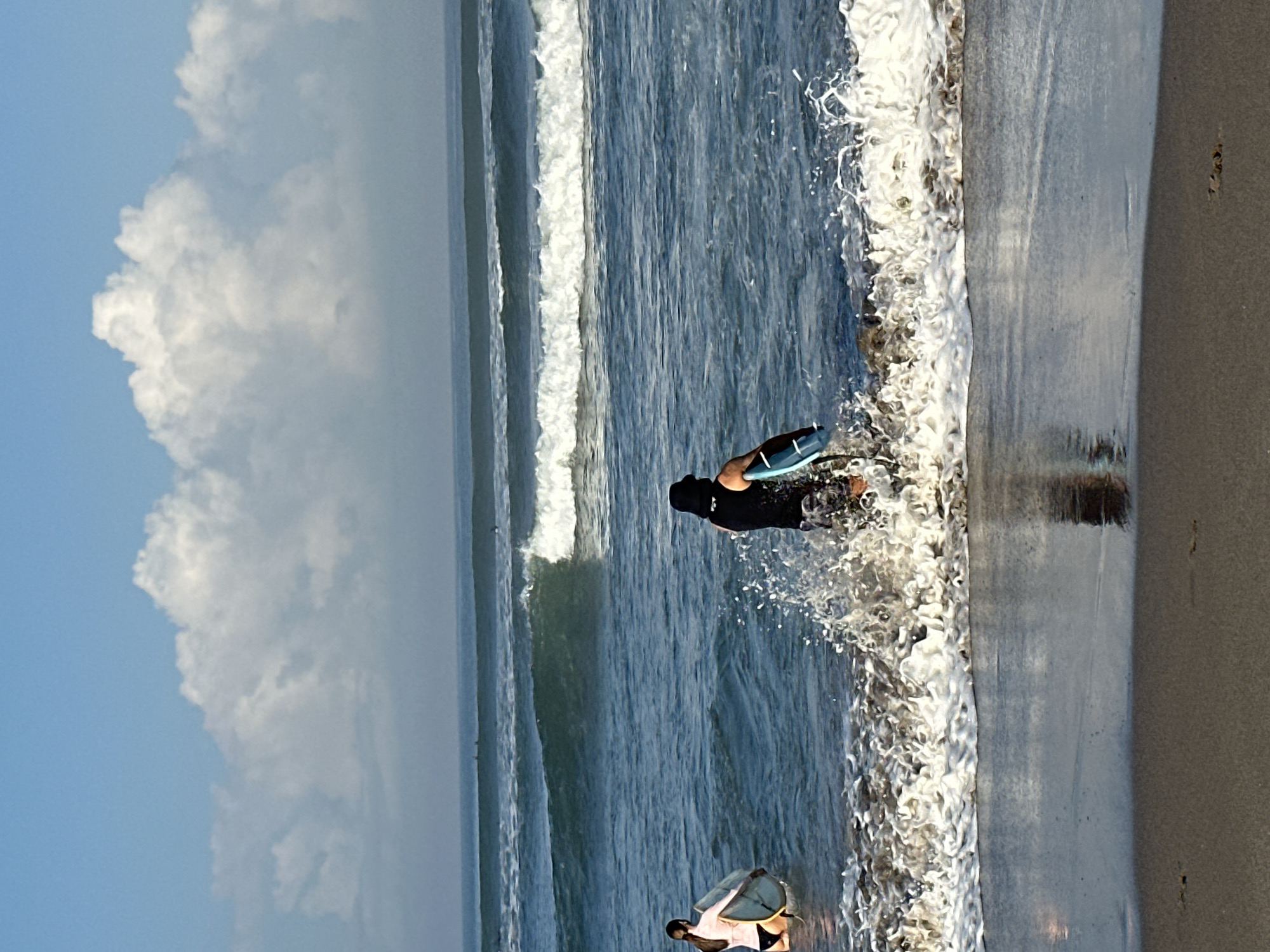 In North Kuta, Indonesia, a sunny day unfolds at the beach with a clear blue sky dotted with fluffy white clouds. The ocean is calm, with gentle waves rolling towards the shore. In the foreground, two individuals are engaged in watersports. A woman, wearing a pink top and black bikini bottoms, stands waist-deep in the water, holding a surfboard. To her right, a person in a black shirt and patterned shorts walks through the crashing waves, carrying a blue surfboard. The wet sand reflects the sky and the ocean, creating a serene beach scene.