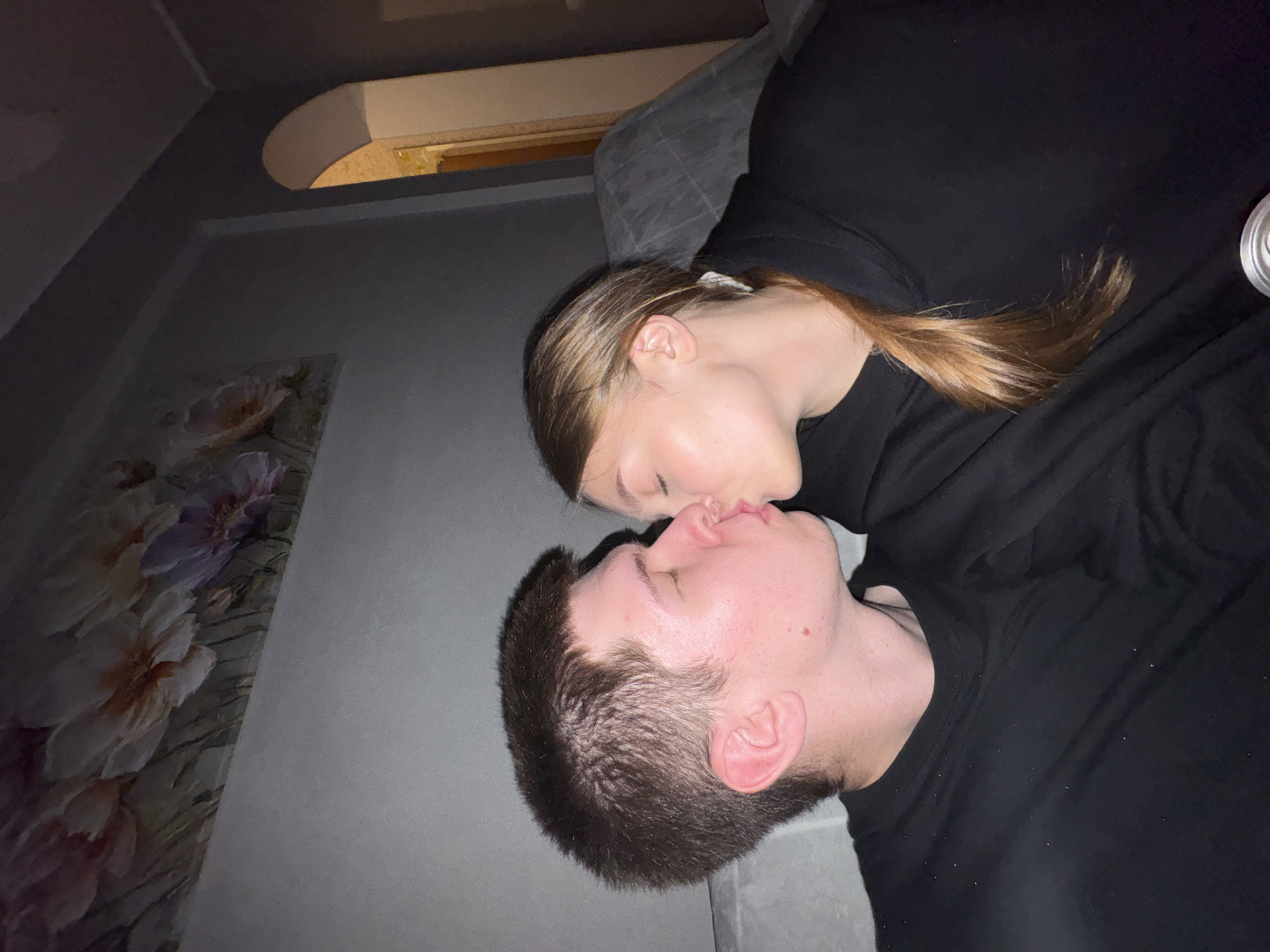 The image depicts two young adults, a male and a female, kissing indoors in a dimly lit residential setting identified as Chaikovsky, Russia. Both individuals have their eyes closed, with their lips pressed together.
The male, positioned on the left, has short, dark brown hair and fair skin, exhibiting a small mole on his right jawline. He is wearing a plain black, long-sleeved crew-neck top, which shows scattered light-colored specks, possibly lint or dust.
The female, on the right, has long, light brown hair pulled back into a low ponytail. She wears a black hooded sweatshirt. Her right ear is visible, and her facial expression mirrors the male's with closed eyes during the kiss.
The background consists of a dark gray wall with a subtle texture. On the upper left portion of the wall, a large painting featuring various flowers in shades of white, pink, purple, and yellow is mounted. To the right, an arched doorway leads to another room, which appears to have a warmer, yellow-toned light source. A dark gray, possibly upholstered, surface of a sofa or chair is partially visible behind the female on the right. In the bottom right corner of the frame, the silver-colored top rim of a metal can is partially visible. The overall lighting is low and ambient, casting soft shadows.