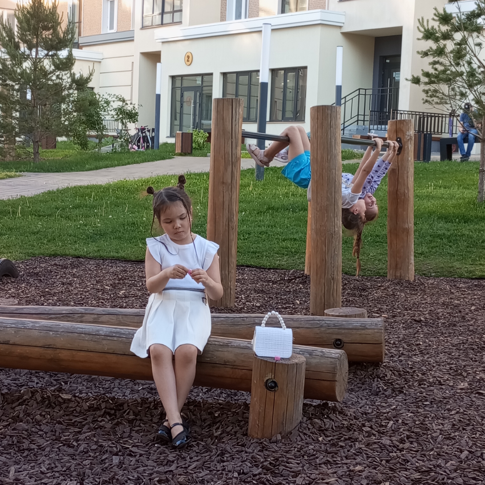 The image depicts a playground in Pervomaysky, Russia, with children engaged in various activities. In the foreground, a young girl with pigtails sits on a wooden beam, holding a small pink object in her hands. She is wearing a white dress and black shoes. Beside her, a white handbag with pearl accents rests on a wooden post. In the background, two other children are on a monkey bar structure, one hanging upside down. The playground features wooden beams, logs, and a soft, wood-chip ground cover. Behind the playground, a modern building with multiple windows and a blue door is visible, along with grassy areas and trees. A person in a blue shirt and jeans sits on a bench to the far right of the frame.