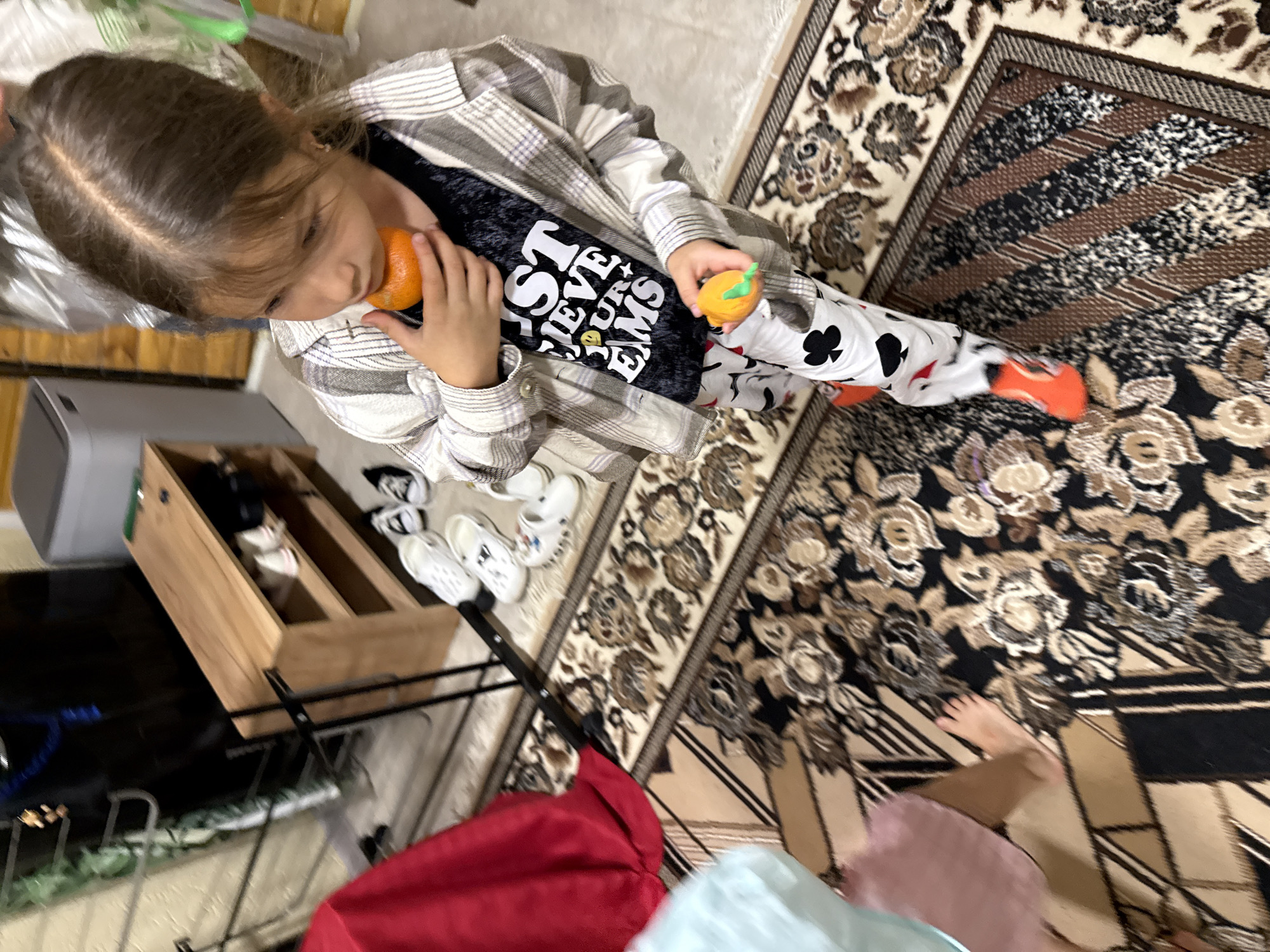 A young girl with brown hair stands on a patterned rug indoors, holding a small orange in her mouth and another in her left hand. She is wearing a plaid shirt over a black t-shirt with white text that reads "JUST BELIEVE YOUR DREAMS" and white pants with a pattern of playing card suits. The room appears to be a hallway or entryway, with a wooden shoe rack holding several pairs of shoes, a red curtain, and a gray appliance visible in the background. The lighting is dim, suggesting it may be nighttime or an interior with limited natural light.