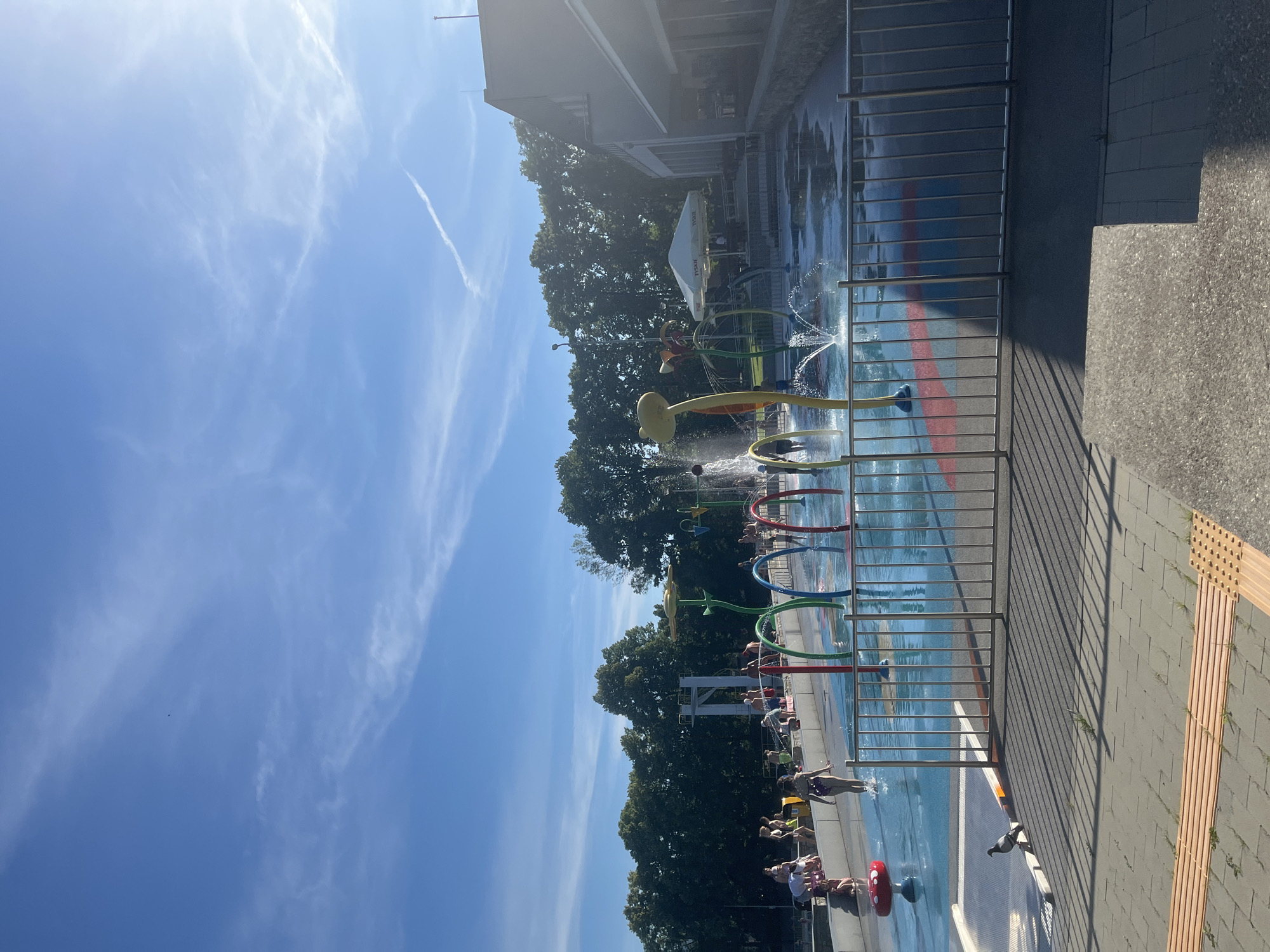 This is a vertical, eye-level, outdoor, daytime photograph of a splash pad. The scene is bright and sunny with a clear blue sky and wispy white clouds. Several people are enjoying the water, with some running through the sprinklers, and others sitting on the edge of the pool. The splash pad features colorful spray features, including large, yellow, mushroom-shaped sprinklers and red and blue hoops that spray water. A metal fence runs along the edge of the splash pad. In the background, there are tall green trees and a building. The ground around the splash pad is paved with gray bricks and wood. The overall atmosphere appears cheerful and fun, suggesting a hot summer day. There are no visible signs or text.