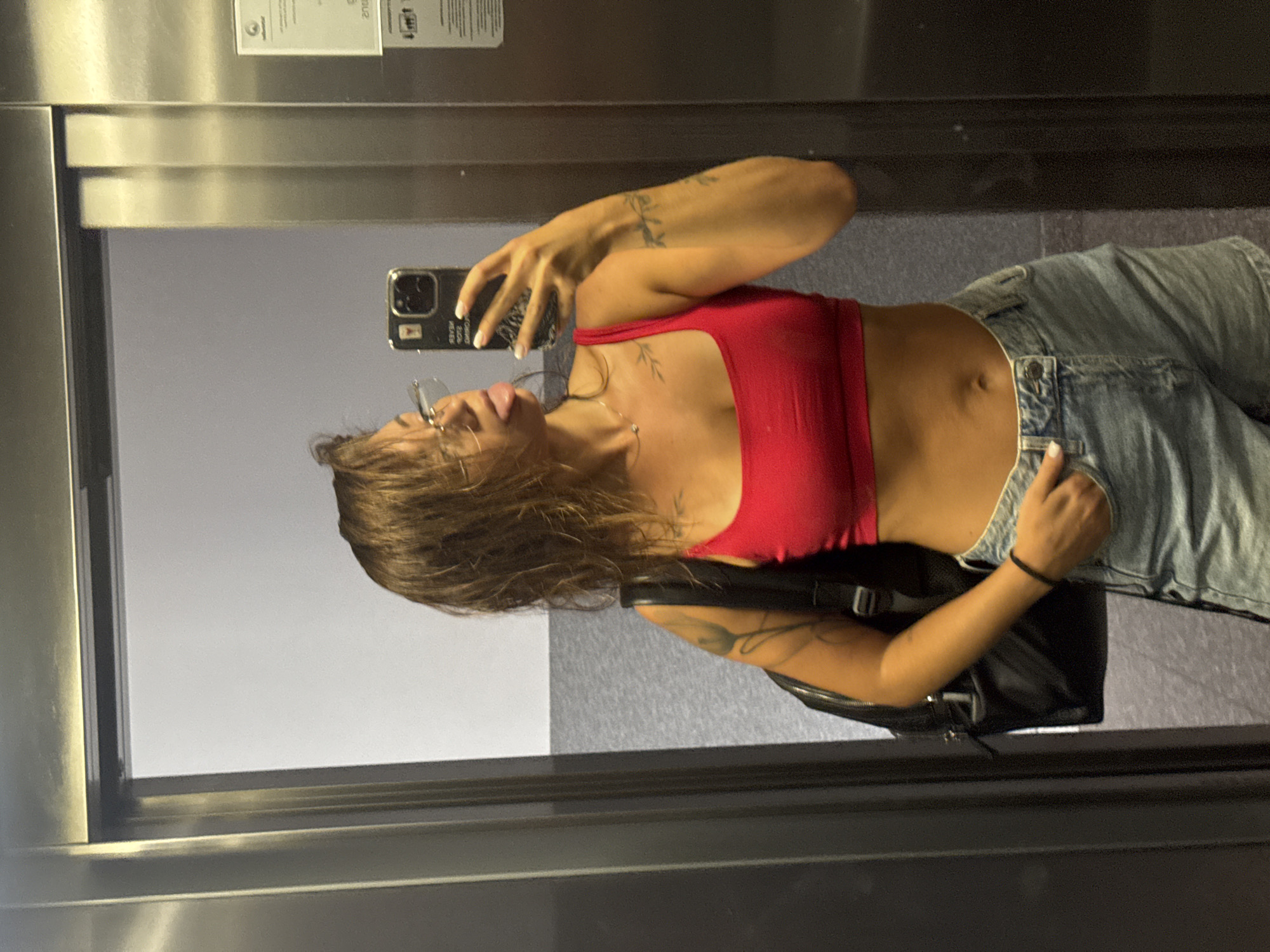 A woman stands in an elevator in Budva, Montenegro, taking a selfie in a mirror. She has medium-length, wet, brown hair and is wearing glasses, a red crop top, and light-wash denim shorts. She is sticking her tongue out at the camera. A black backpack is slung over her left shoulder. The elevator walls are metallic, and the floor appears to be tiled. A sign with text and a logo is visible on the right-hand elevator door.