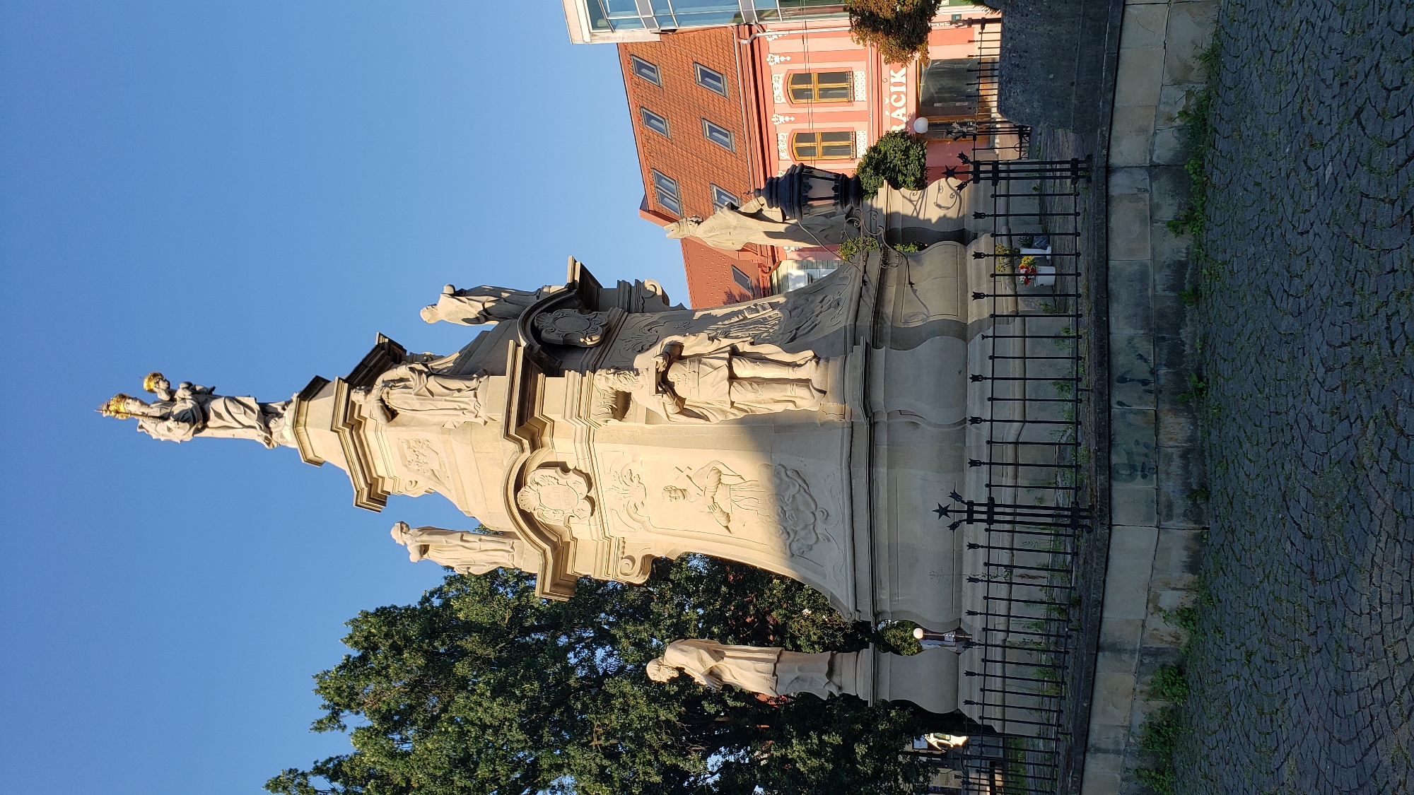 The image shows the Holy Trinity Column located in the city of Prešov, Slovakia. The column, constructed from light-colored stone, is a prominent baroque monument. It is adorned with numerous statues and reliefs depicting religious figures. At the very top, a statue of the Virgin Mary holding the infant Jesus is visible, both wearing golden crowns. The column is surrounded by a low black metal fence and sits on a cobblestone plaza. In the background, buildings with red-tiled roofs and colorful facades are visible, along with a large tree on the left side of the frame. The sky is clear and blue, suggesting it is daytime.
