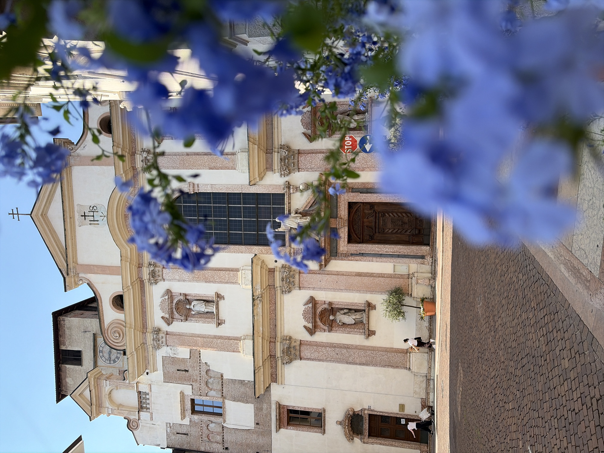 The image shows a religious building, likely a church, with a prominent facade. The building is made of stone and features architectural details like columns, arches, and statues. A clock is visible on one side of the building. In the foreground, blue flowers are blurred, suggesting they are part of a decorative element or hanging plants.
The scene appears to be a public space, possibly a plaza or street, paved with cobblestones. A few people are present, walking along the paved area. One person is wearing a white mask, indicating a contemporary setting. A stop sign and a directional sign are visible on the building's facade. The sky is clear, suggesting fair weather, and the lighting indicates it is daytime, possibly late afternoon given the shadows. The architectural style and the presence of a church suggest a European location, consistent with the provided context of Trento, Italy. The overall mood is serene and contemplative, with a touch of vibrancy from the flowers.